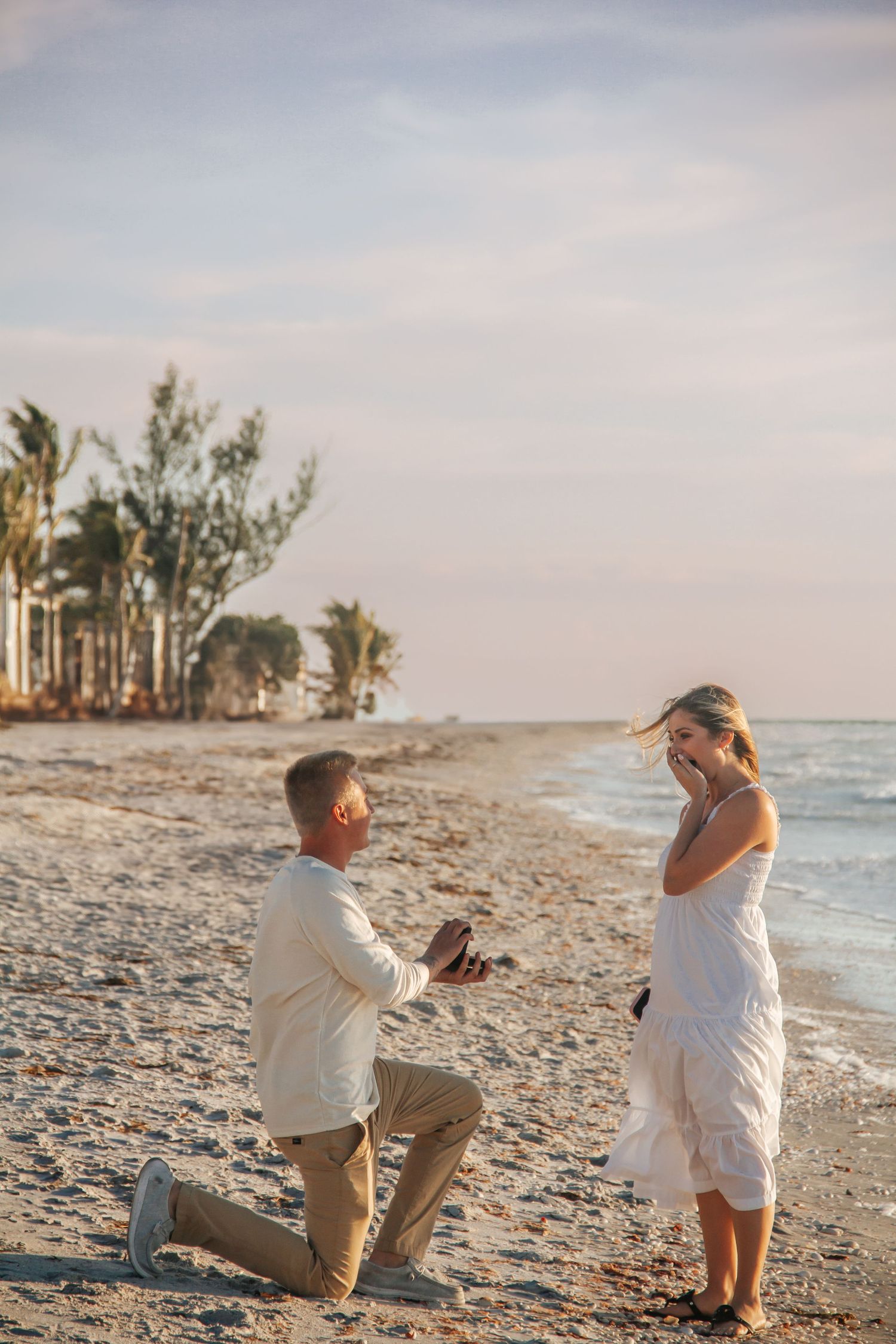 Man proposes on bended knee to woman in white dress on a sandy beach at sunset with palm trees.
