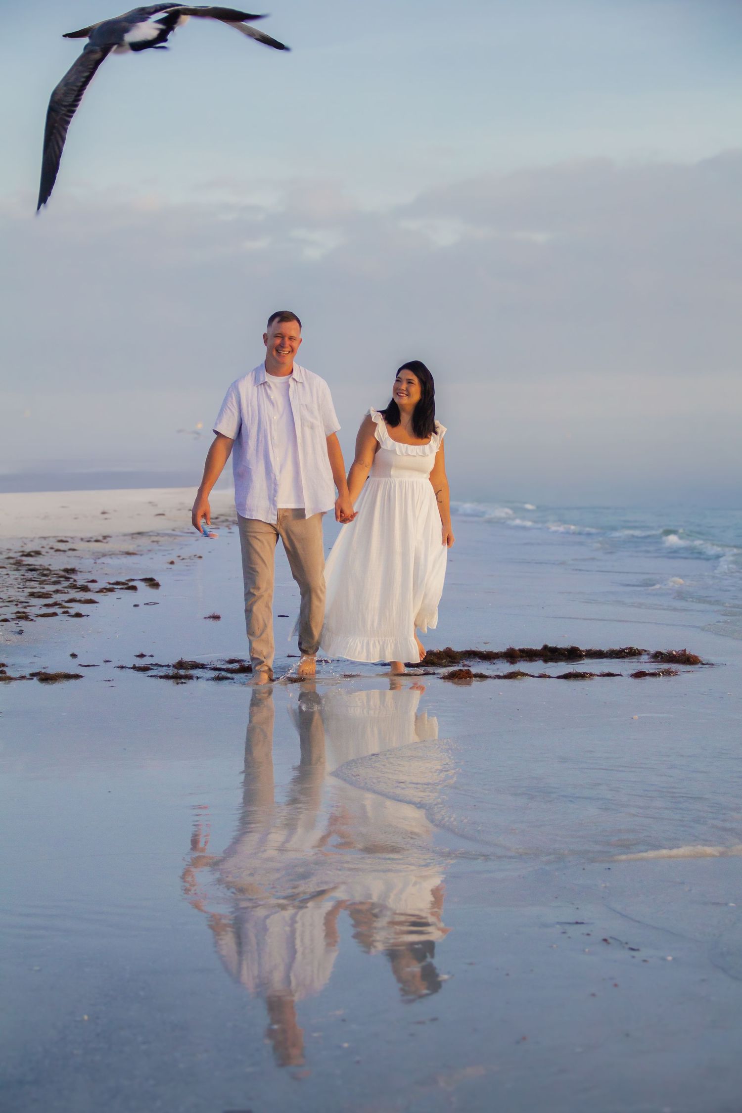 Couple in white attire holding hands walking barefoot on a reflective beach with a seagull flying overhead.