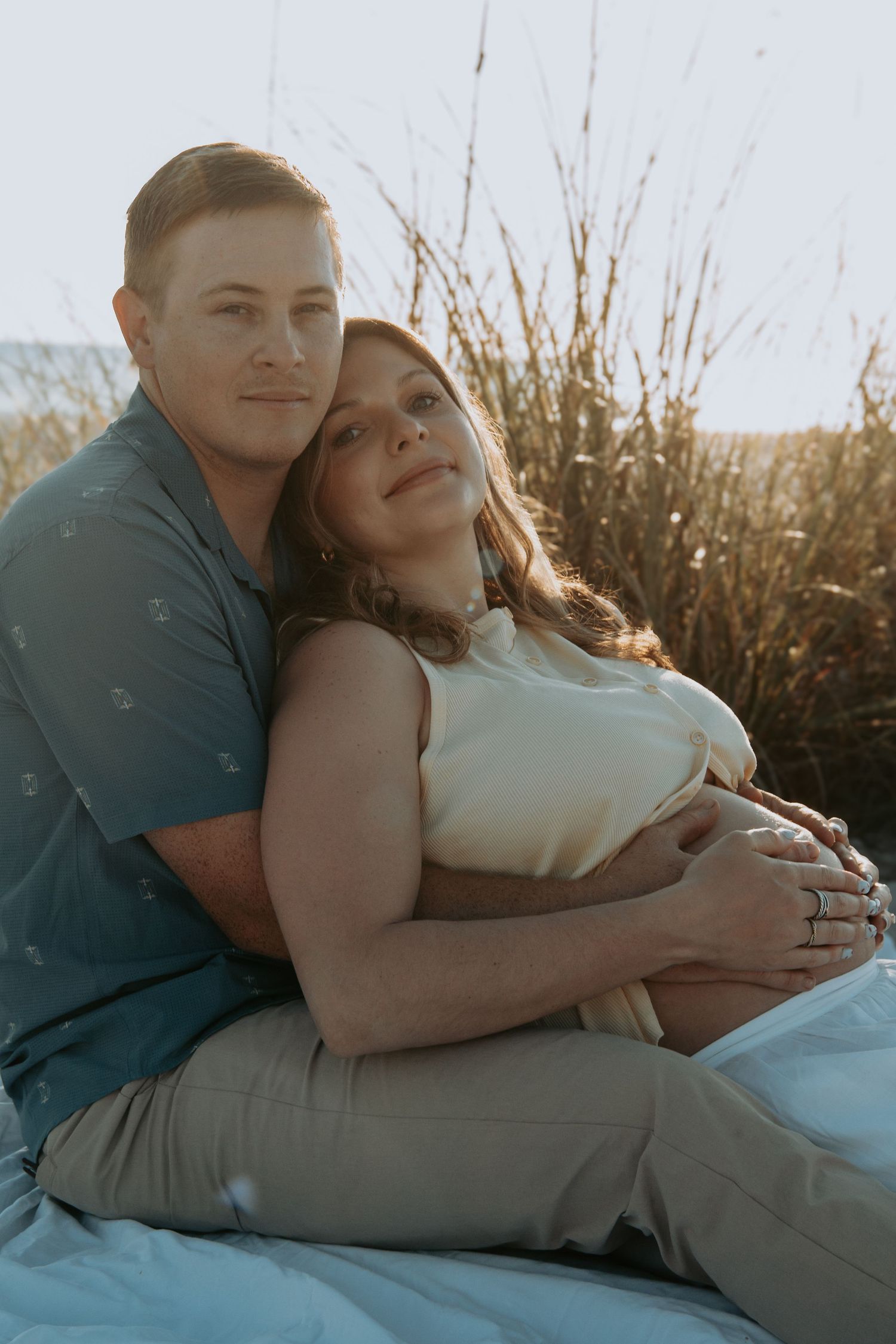 A couple sits outdoors at golden hour, the man embracing a pregnant woman from behind near tall grass by the water.