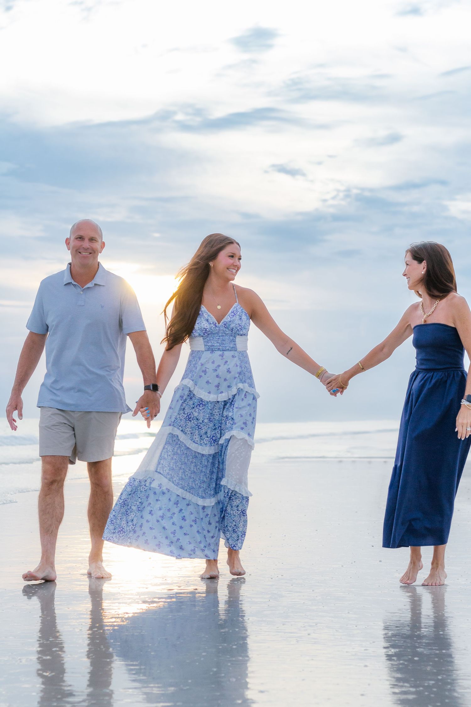 Three people holding hands and walking on a reflective beach at sunset, wearing blue outfits, smiling joyfully.