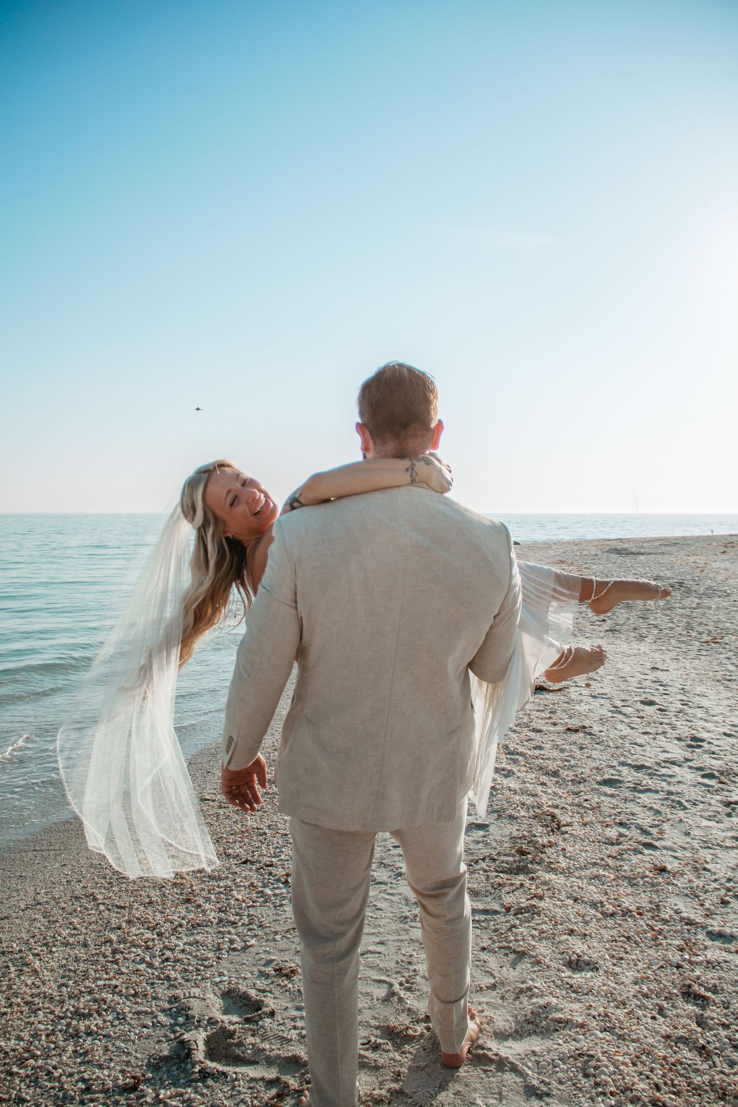 Groom carrying bride in white veil on sandy beach with calm ocean backdrop on their wedding day.
