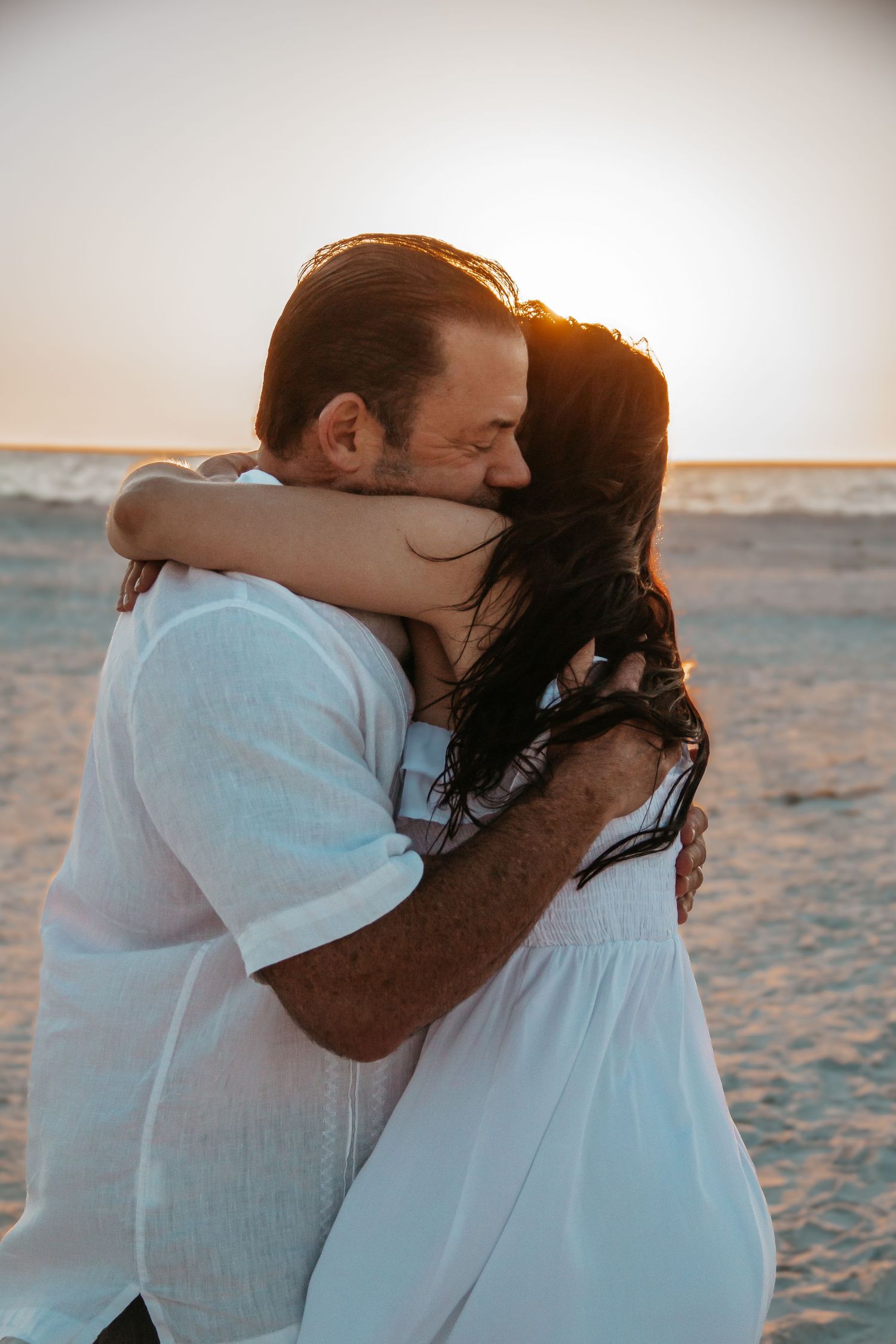 Couple embracing on beach at golden sunset, woman in white dress with dark hair, man in white shirt.