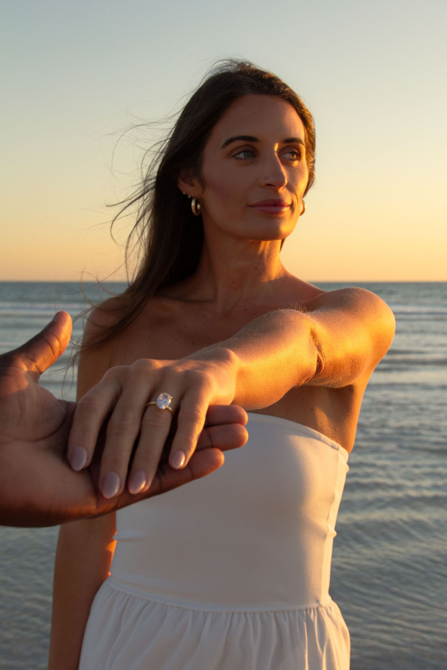 Woman showing diamond engagement ring on beach at golden hour sunset in white strapless dress.