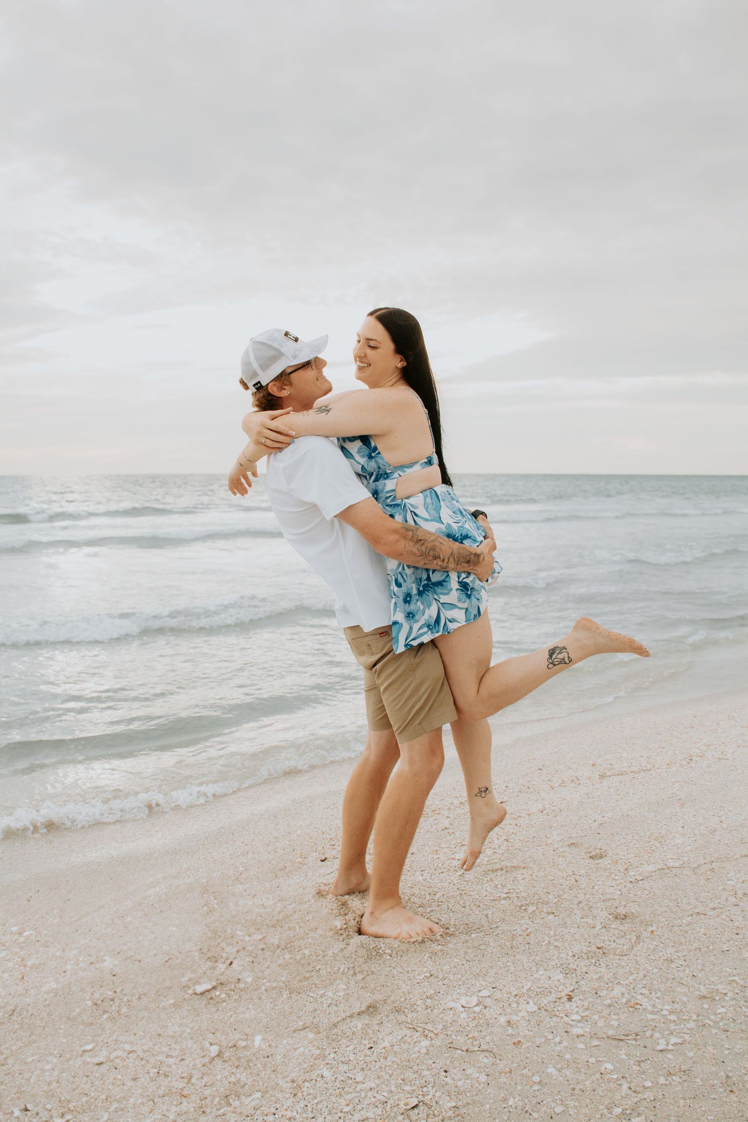 A couple embraces joyfully on a beach, the man lifting the woman as waves roll in behind them.