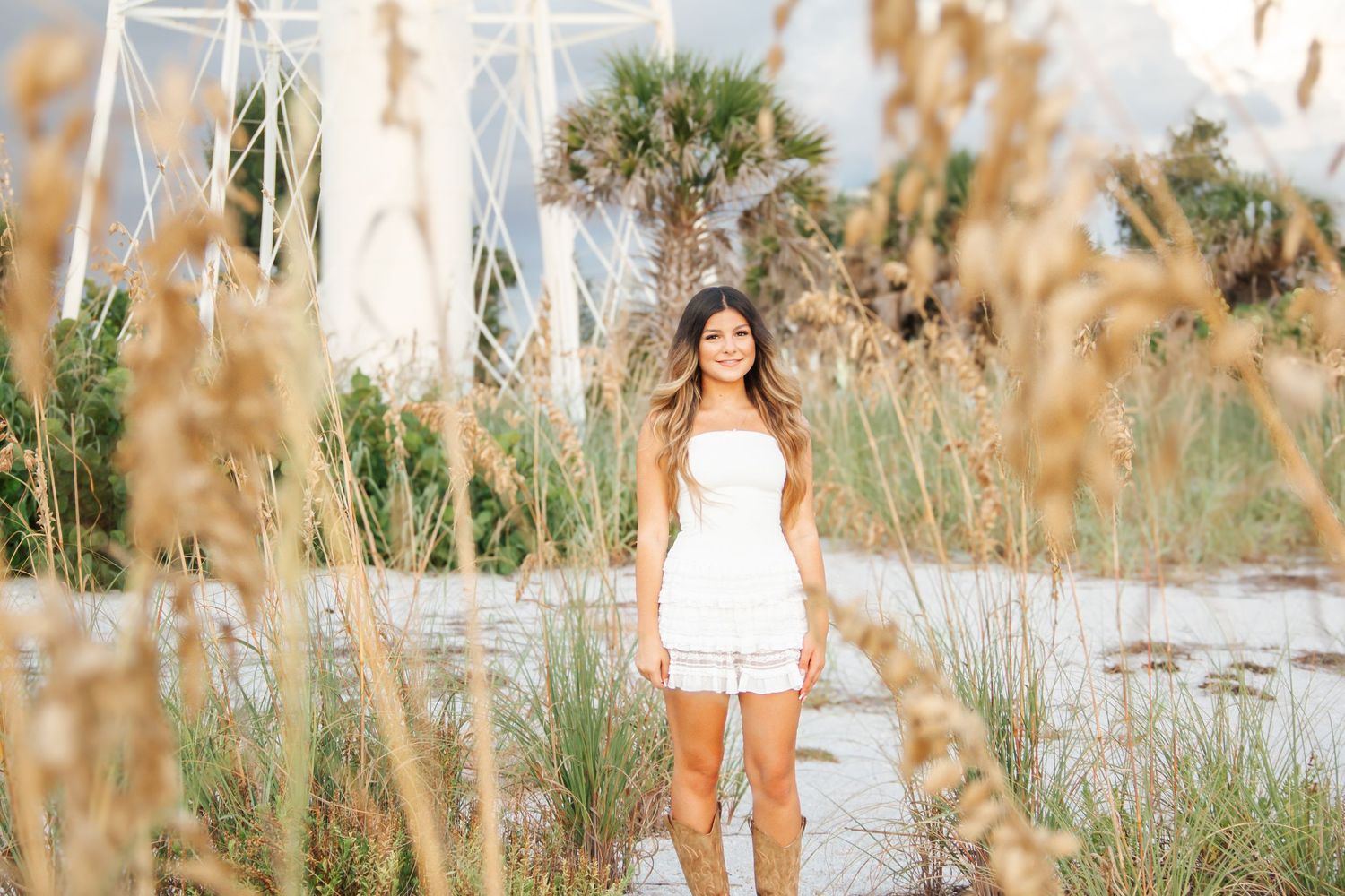 Young woman in white dress standing among sea oats on a sunny beach with palm trees in background.
