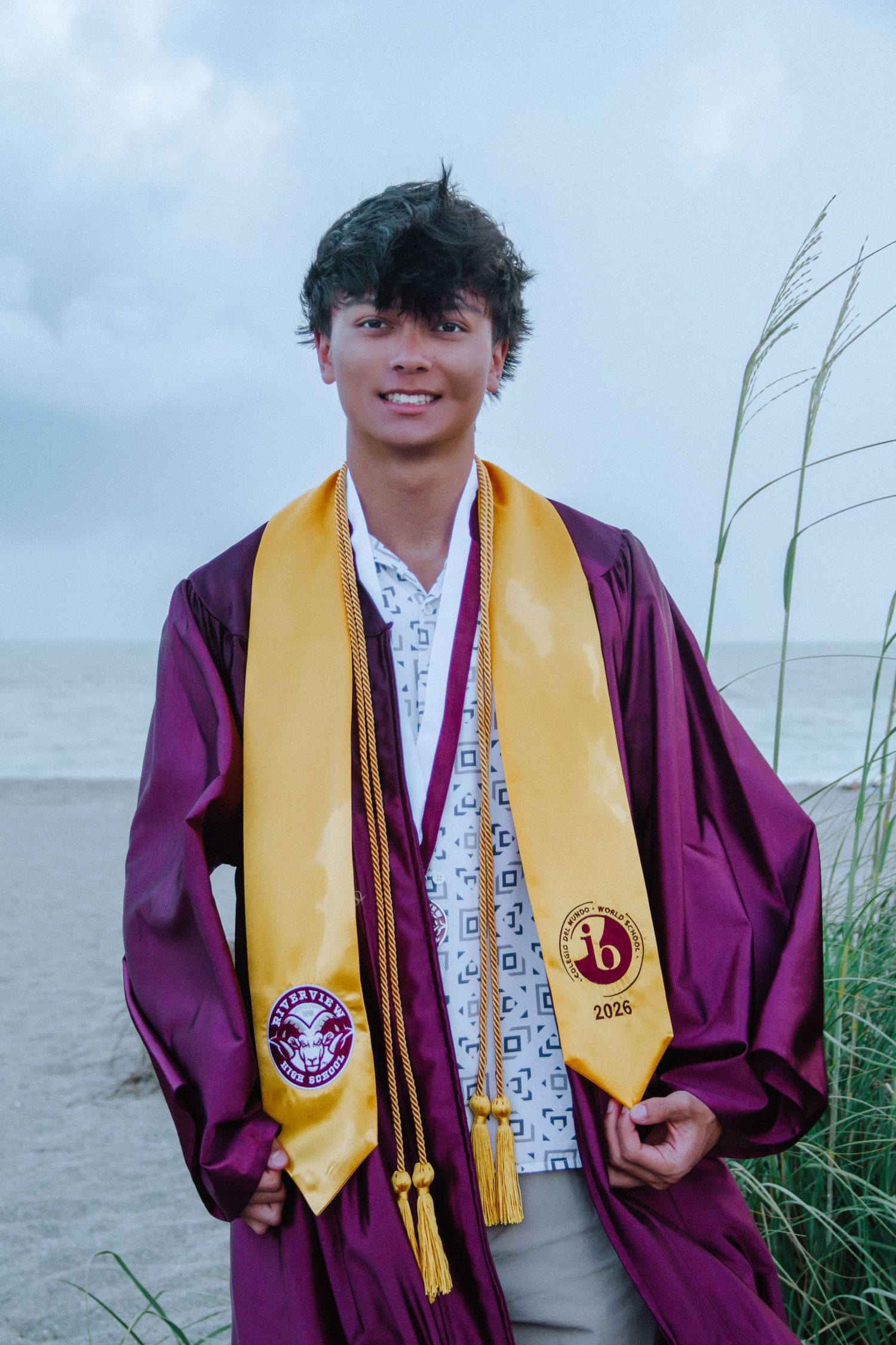 Smiling young man in maroon graduation gown and gold stole poses on beach with tall grass backdrop.