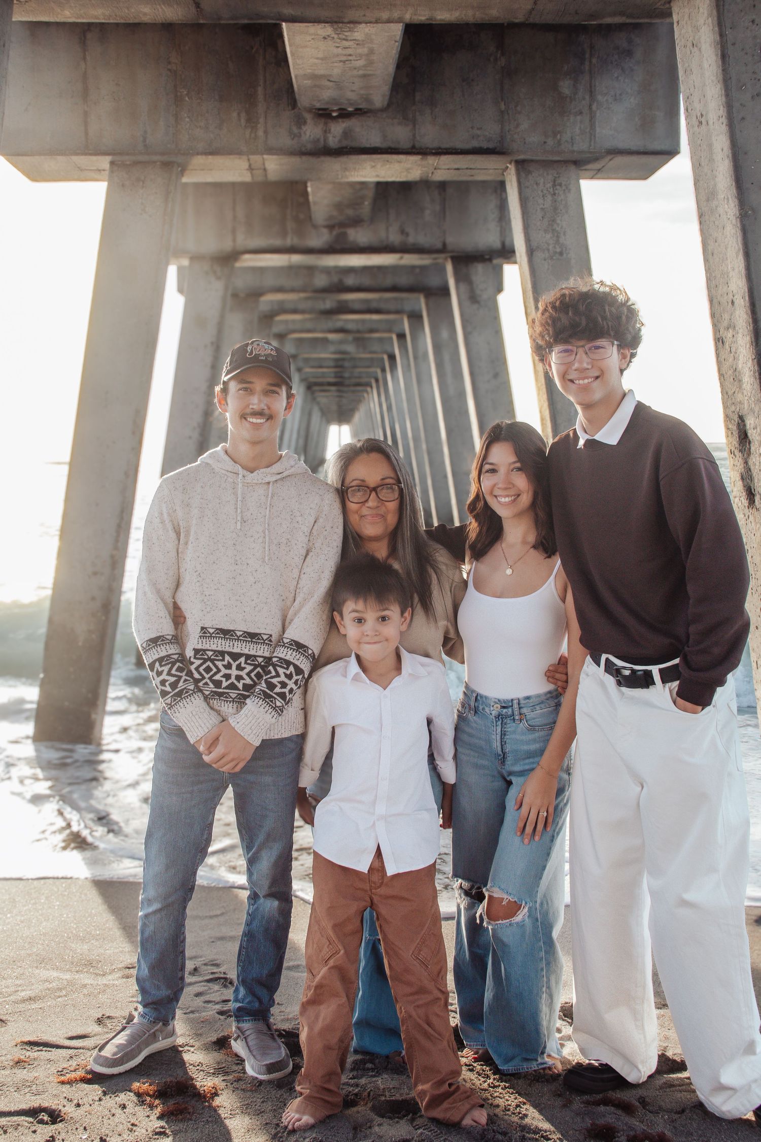 Family of five posing under a beach pier, smiling together with ocean waves and sunlight in the background.