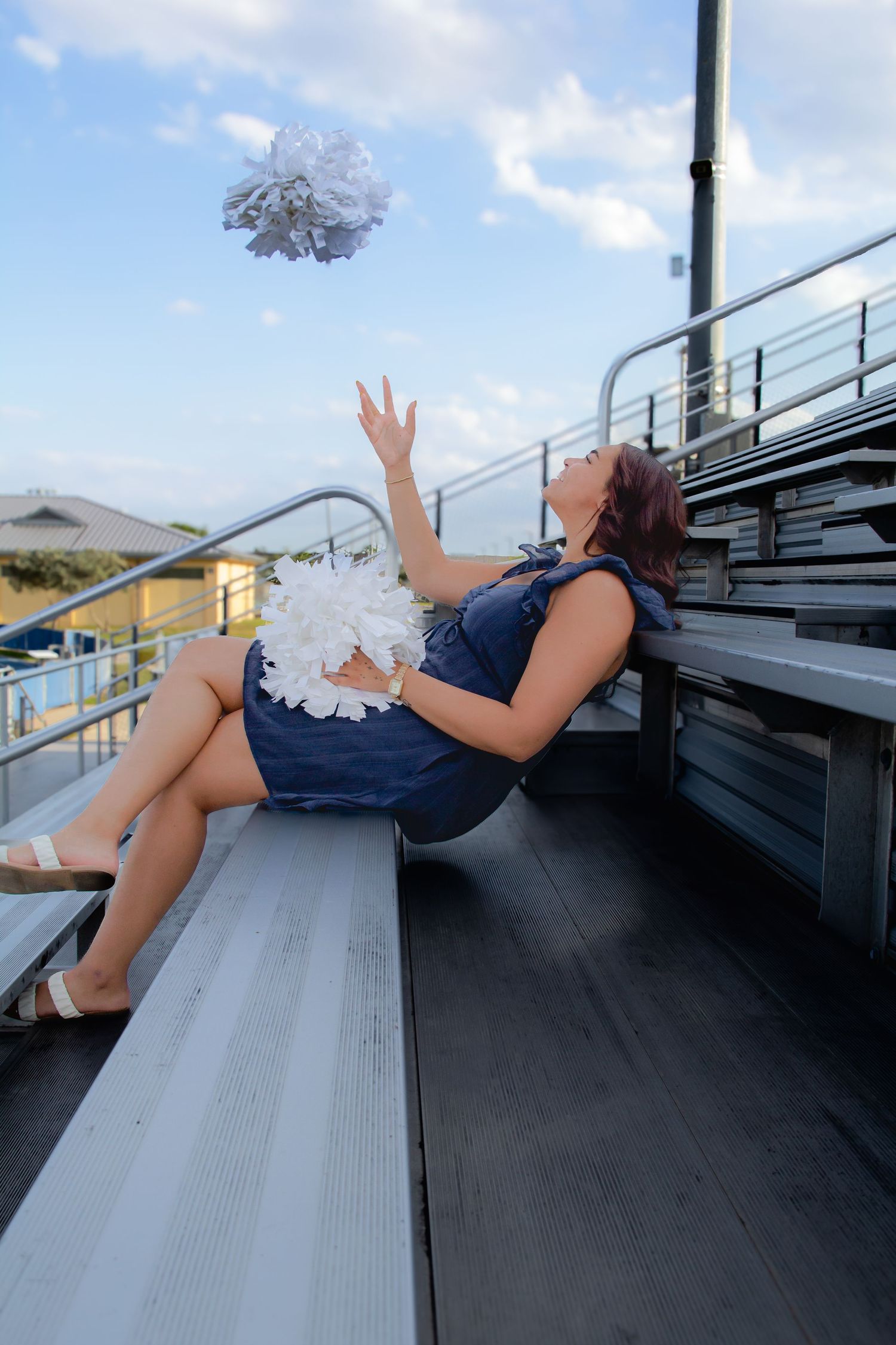 A cheerleader in a blue dress tosses a white pom-pom in the air while sitting on bleachers outdoors.