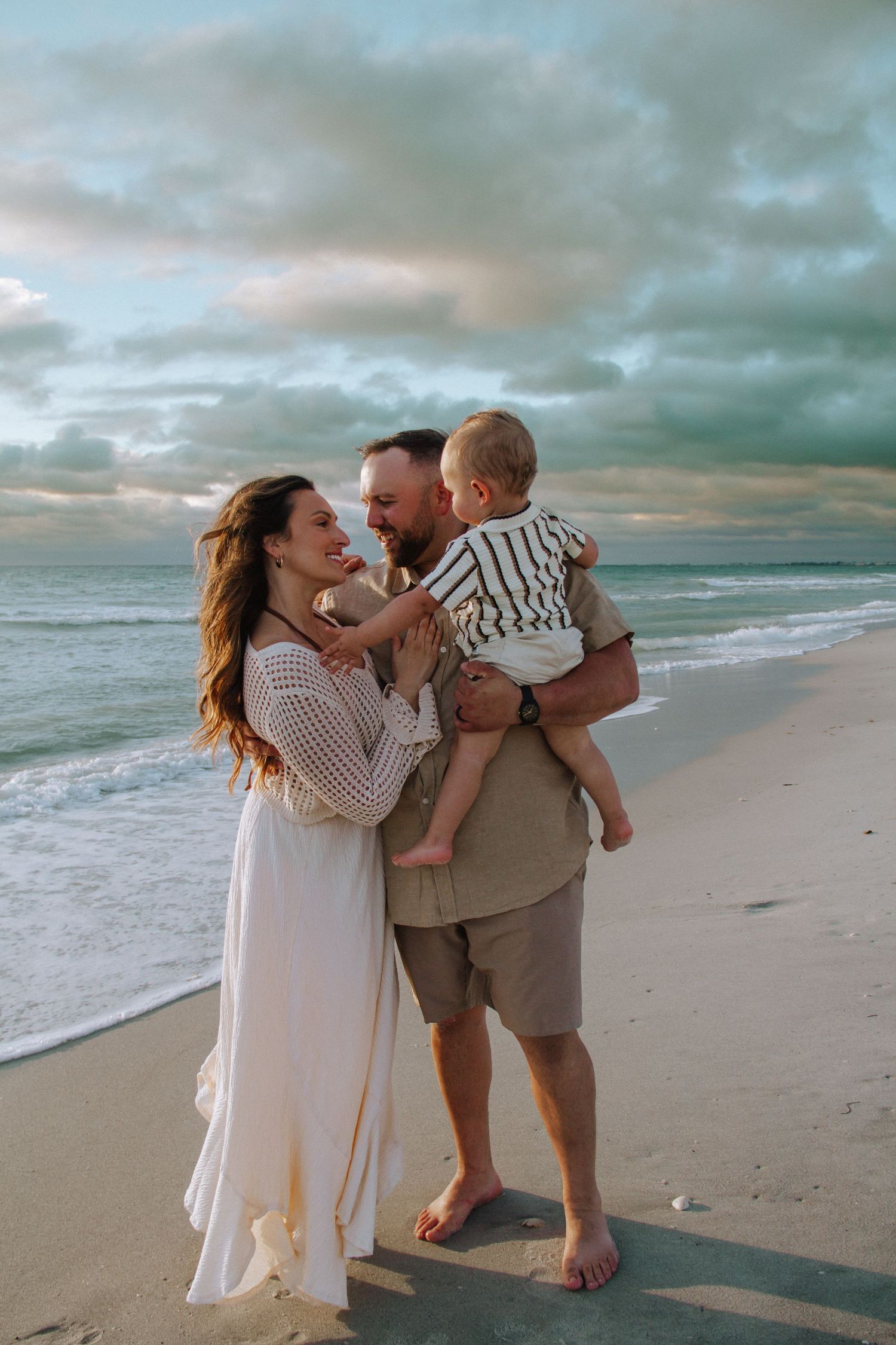 A happy family of three enjoys a sunset beach stroll, with parents smiling at their toddler on a cloudy shore.