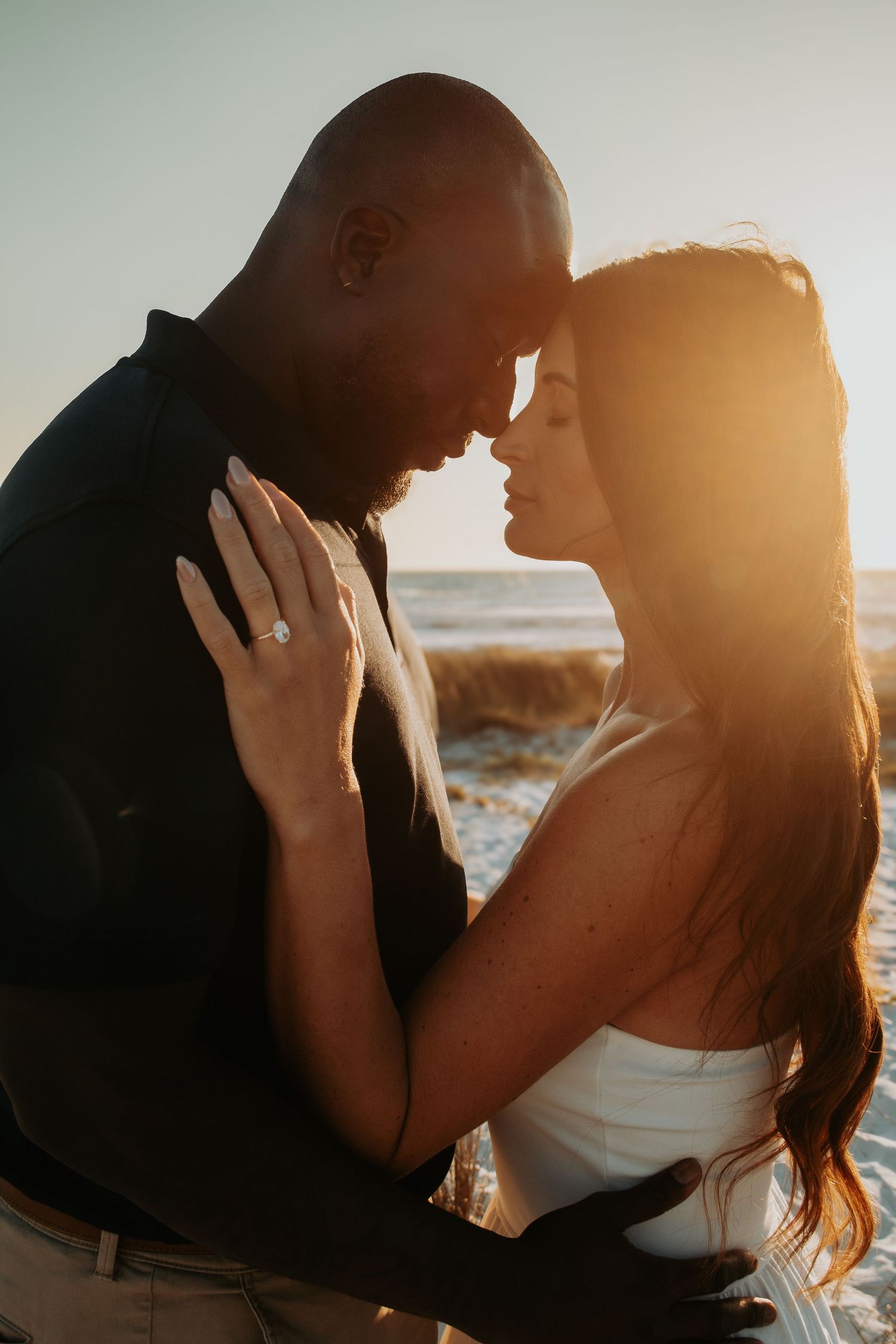Couple sharing a romantic sunset moment on the beach, woman wearing an engagement ring, golden backlight glowing behind them.
