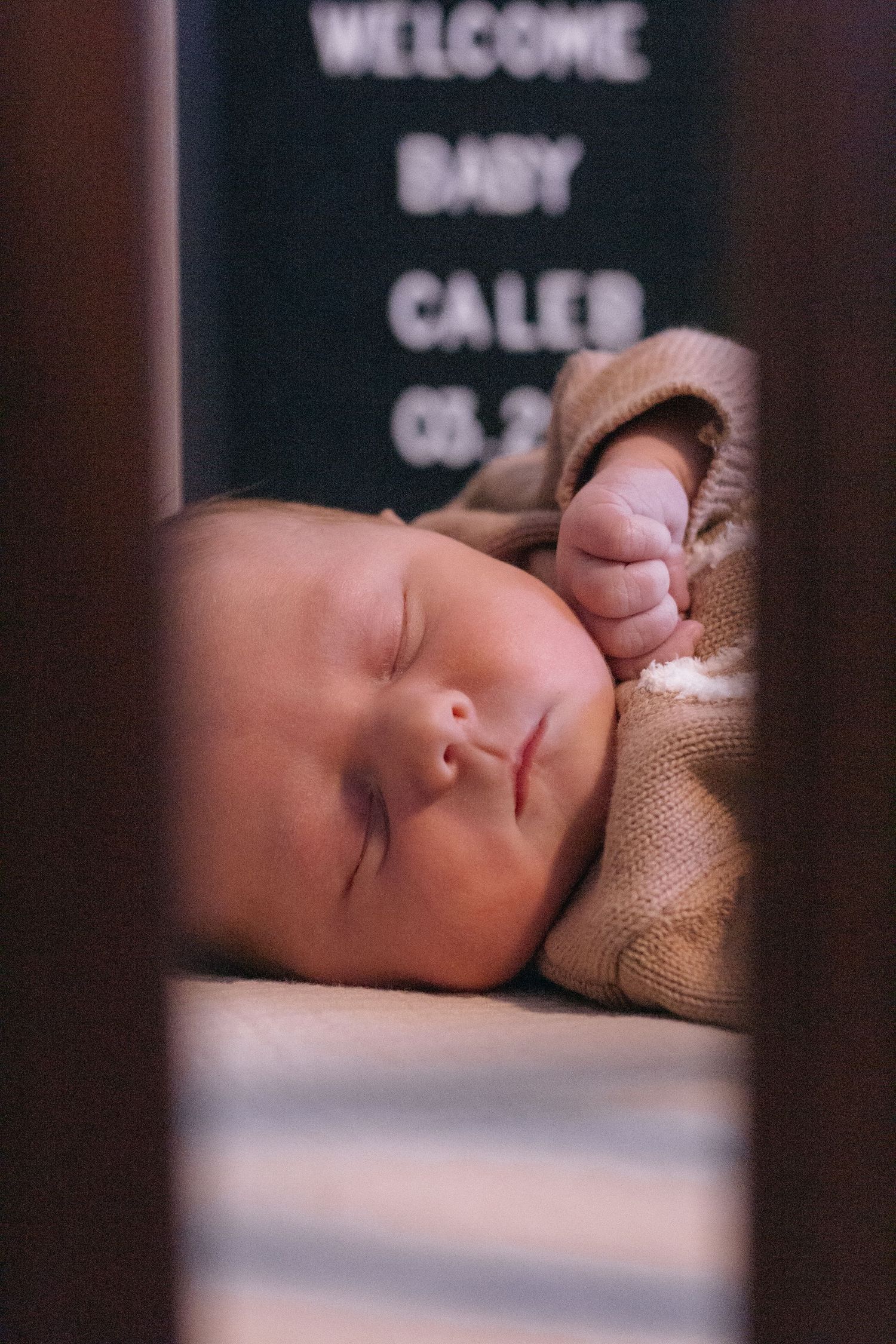 Newborn baby Caleb sleeping peacefully in a crib, wearing a tan knit outfit, with a welcome sign visible in the background.