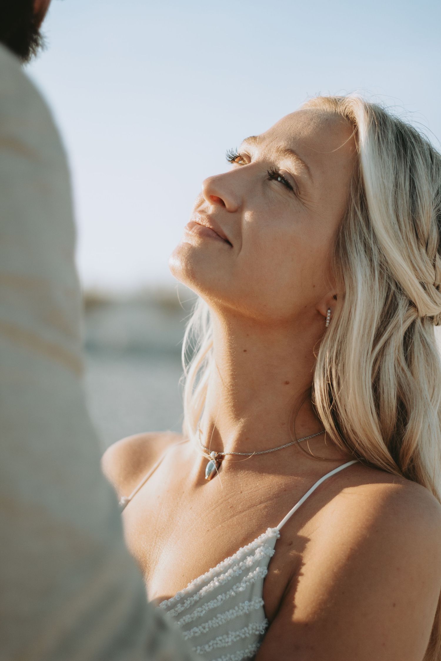 Blonde woman in white dress gazes up at golden hour light outdoors, wearing a delicate pendant necklace.