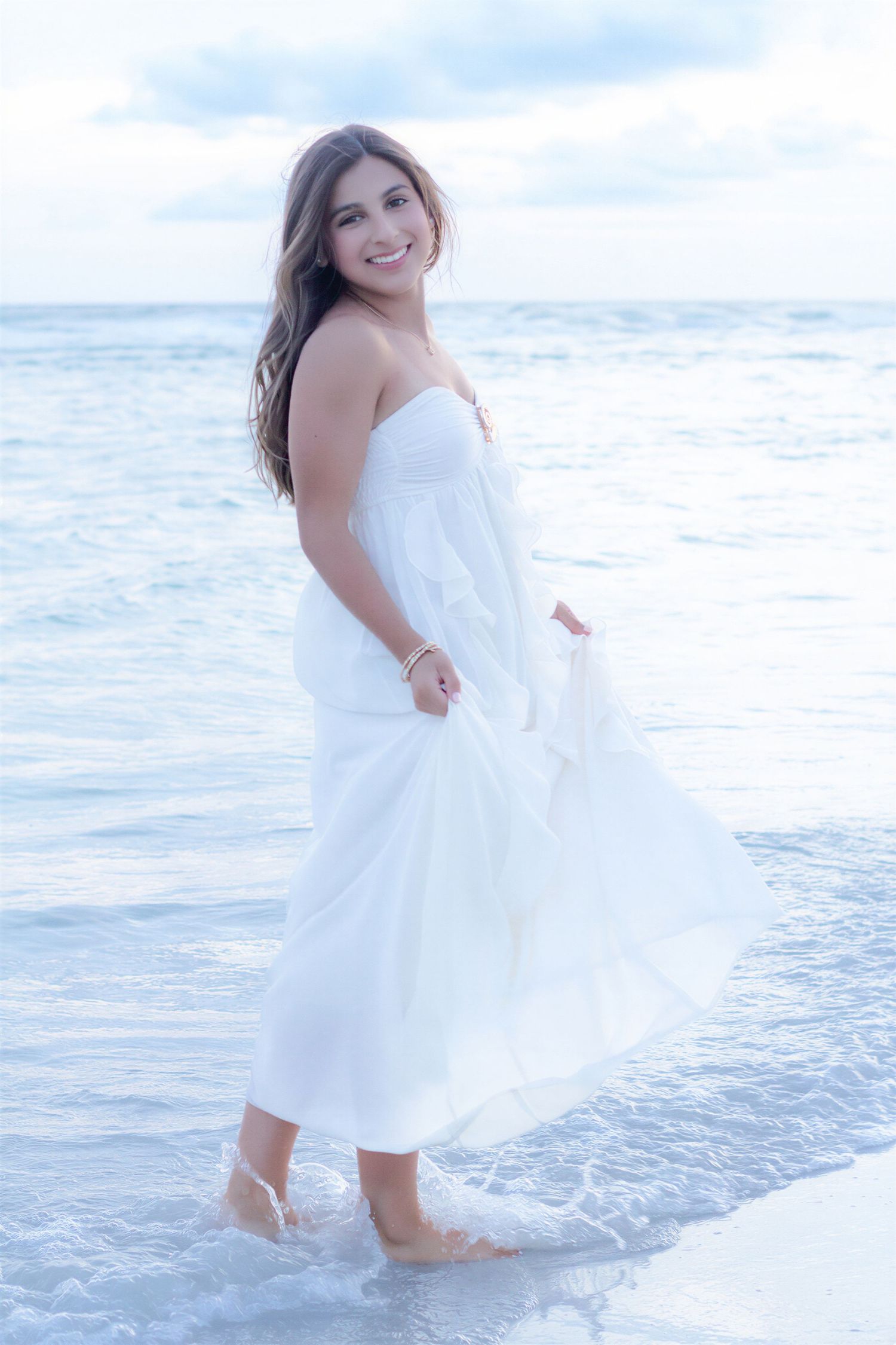 Smiling woman in white strapless dress standing barefoot in ocean waves on a beach.