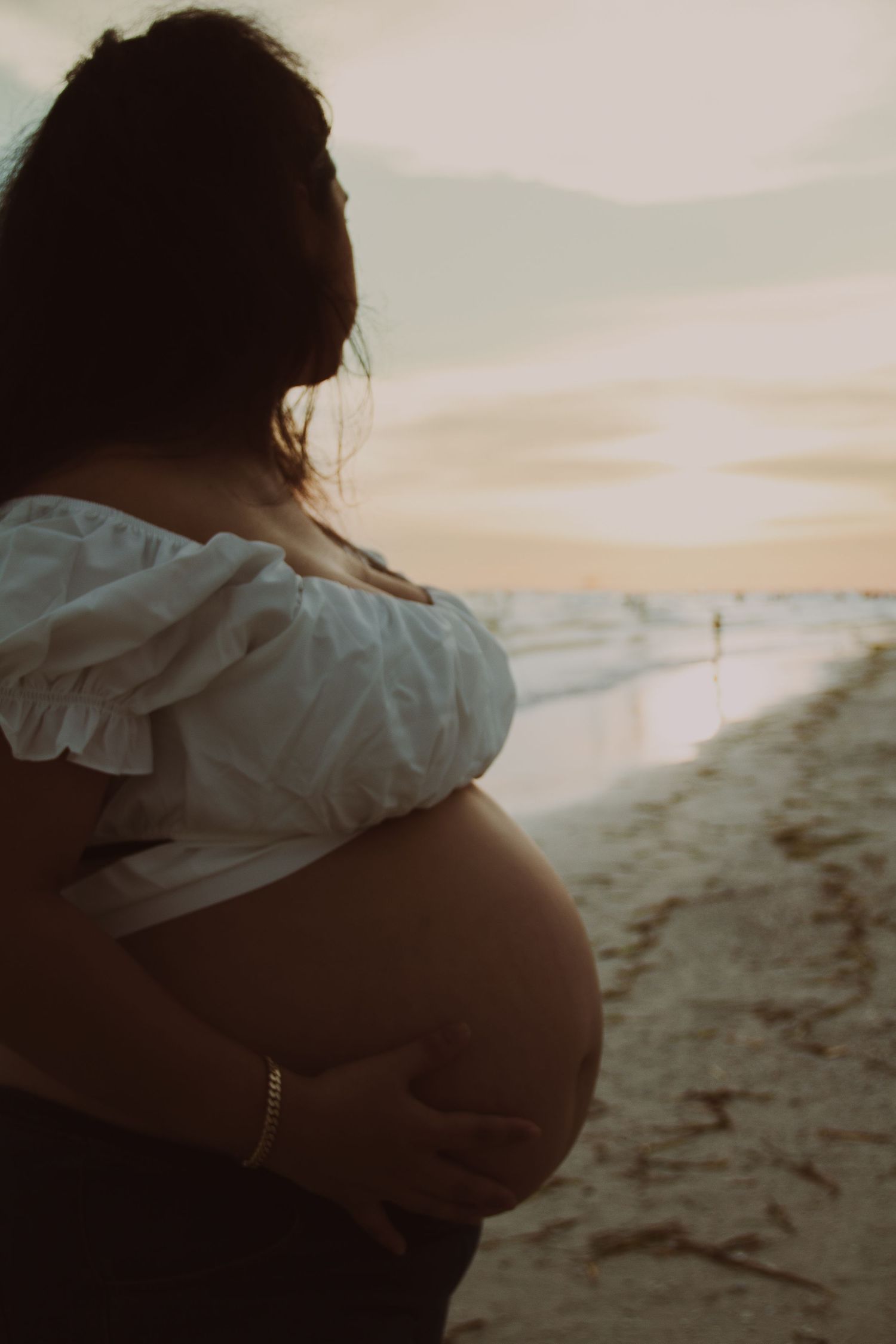 Pregnant woman in white off-shoulder top cradling her belly on a beach at sunset.