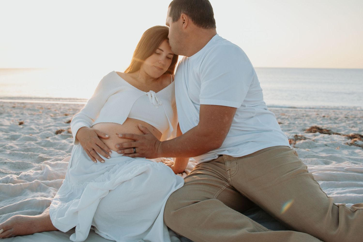 Couple sharing a tender moment on a sandy beach at sunset, with man kissing pregnant woman's head.