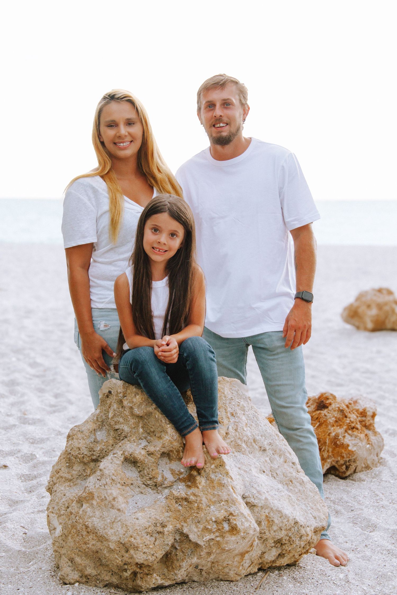 Happy family of three posing on rocks at the beach, wearing white shirts and jeans, smiling at the camera.