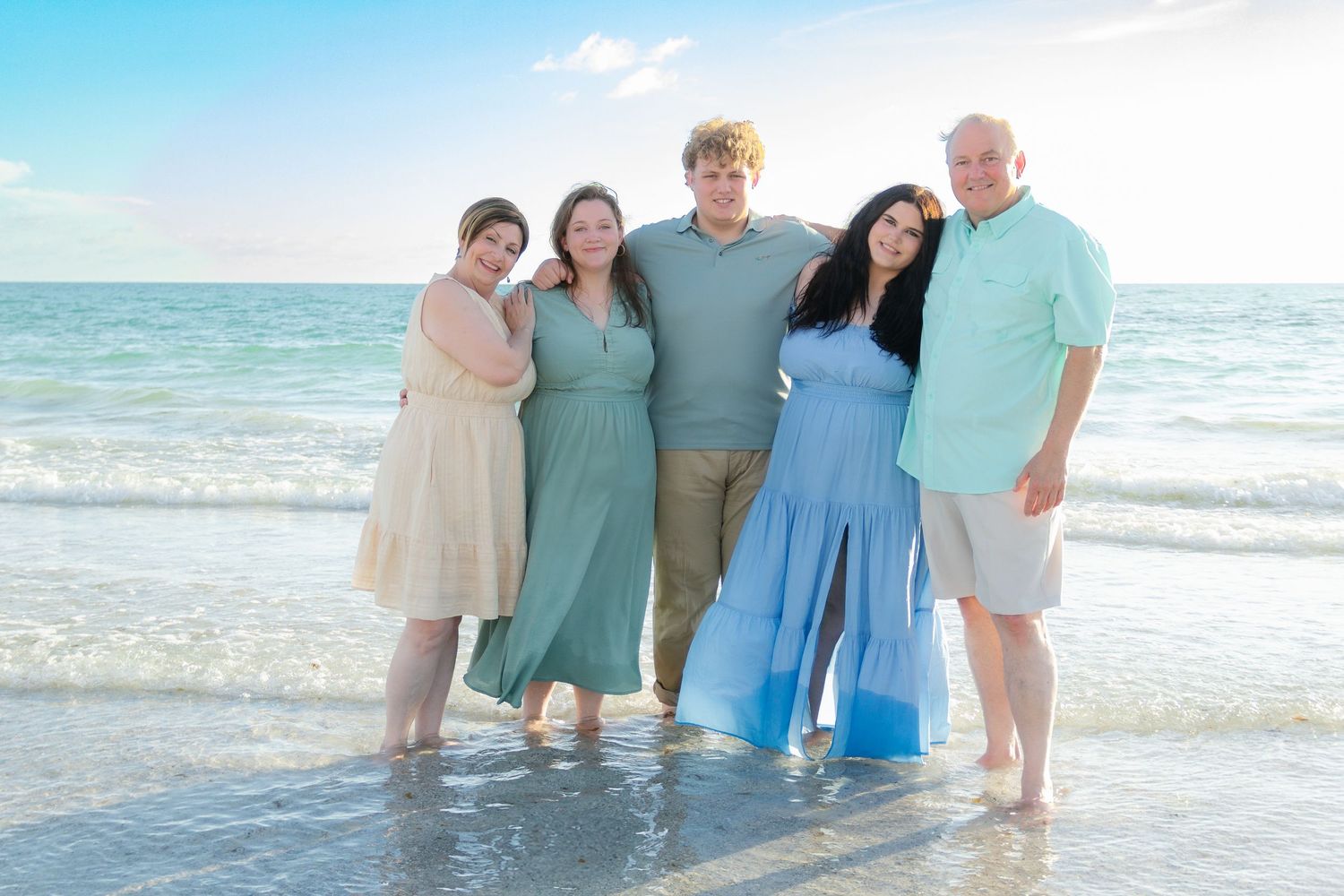 Family of five posing together on a beach at sunset, wearing coordinated pastel outfits with ocean waves behind them.