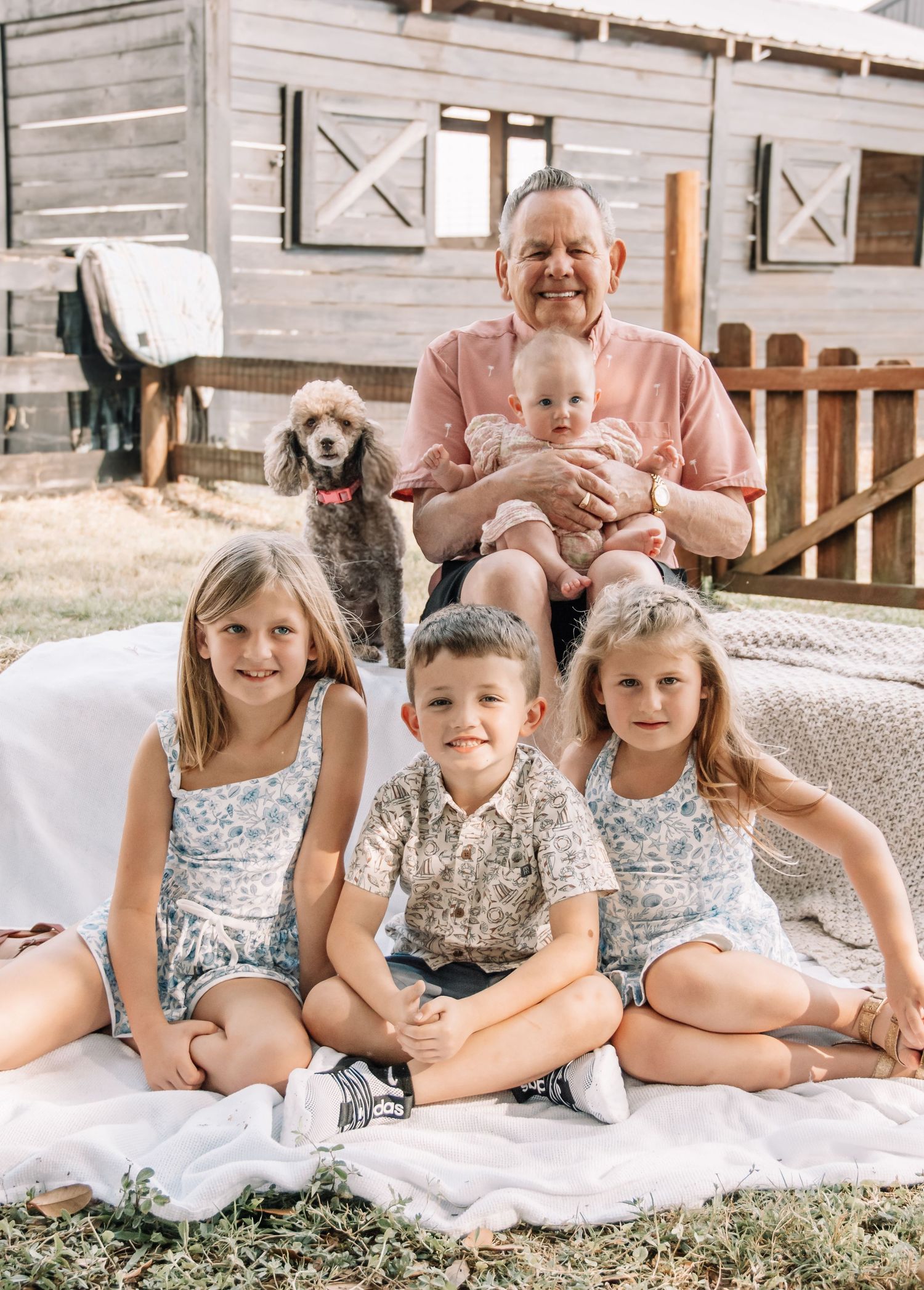 Grandfather smiling with four grandchildren and a gray poodle outdoors near a rustic wooden barn on a sunny day.