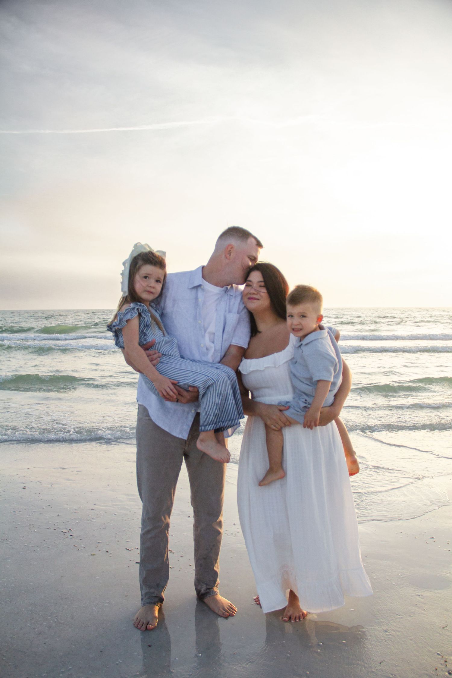 Family of four posing on a beach at sunset, parents holding two young children in coordinating blue and white outfits.