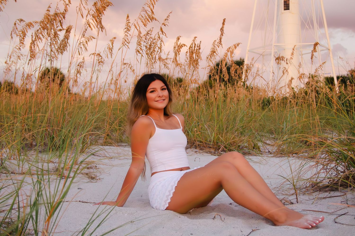 Young woman in white outfit sitting on sandy beach dunes with sea oats and lighthouse at golden hour.