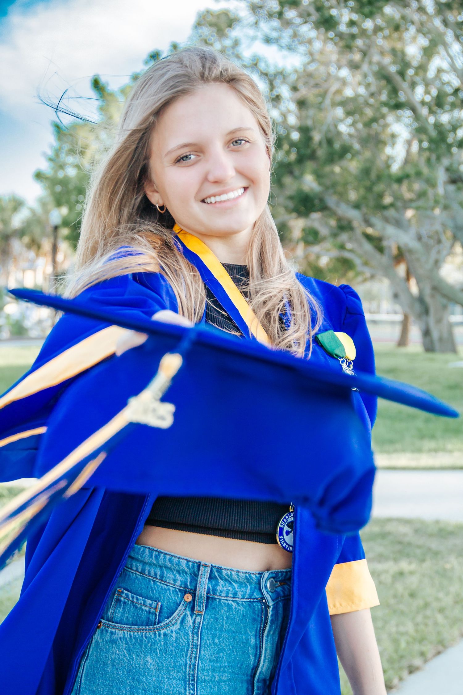 Smiling young woman in blue graduation gown and cap tossing mortarboard outdoors on a sunny day.