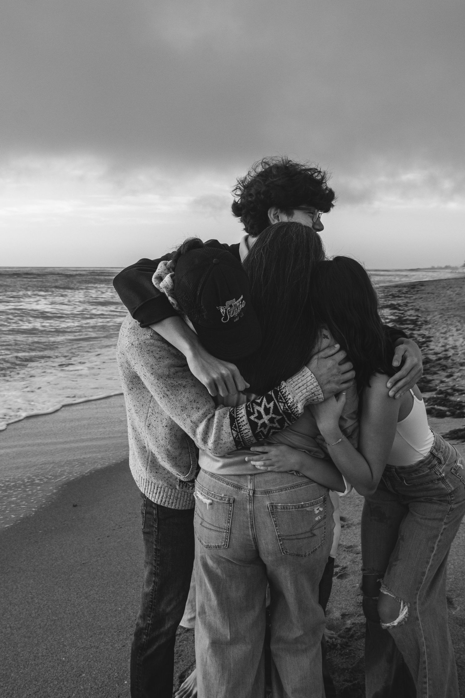 Group of friends sharing a warm embrace on a sandy beach with dramatic cloudy skies in black and white.