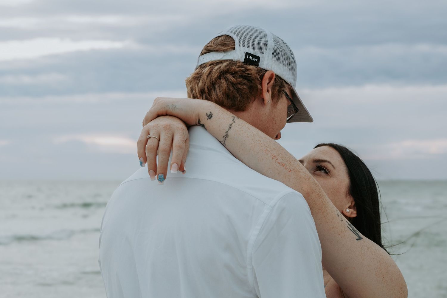 Couple embracing on a beach, woman with dark hair resting her head on man's shoulder near the ocean.