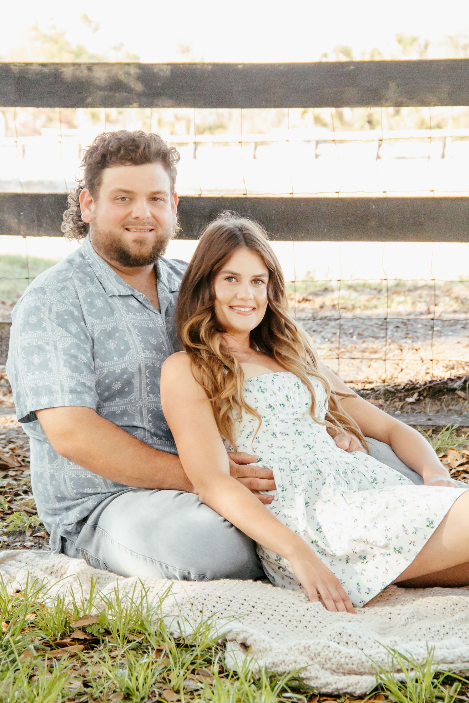 Couple sitting on a blanket by a wooden fence in golden hour light, woman in floral dress, man in blue shirt.