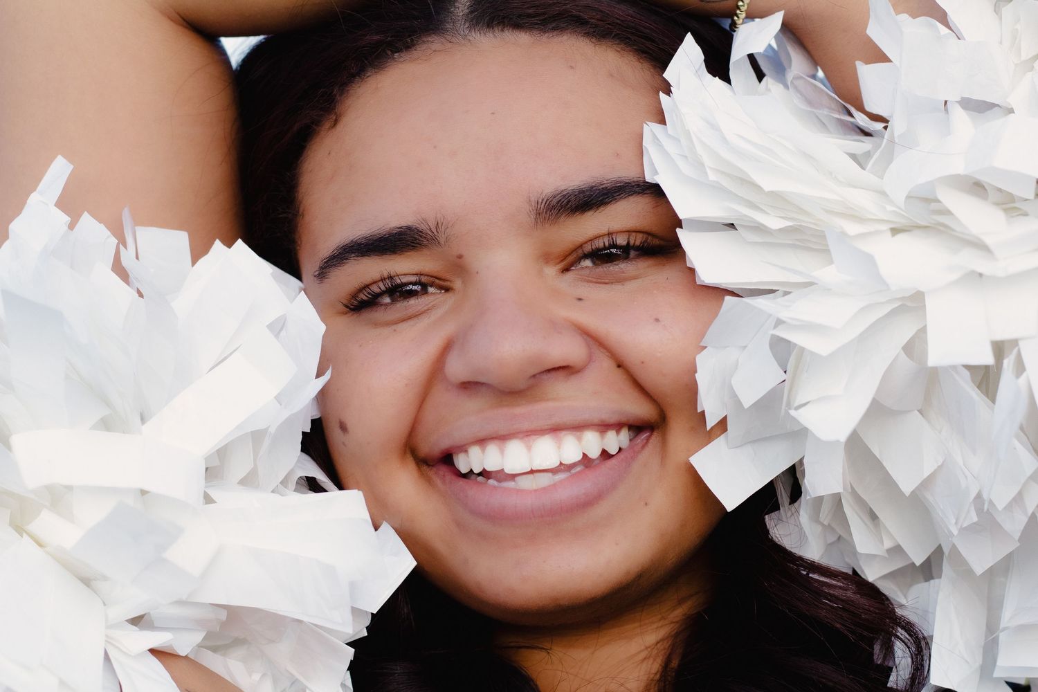 Smiling cheerleader holding white pom-poms close to her face, radiating joy and team spirit.