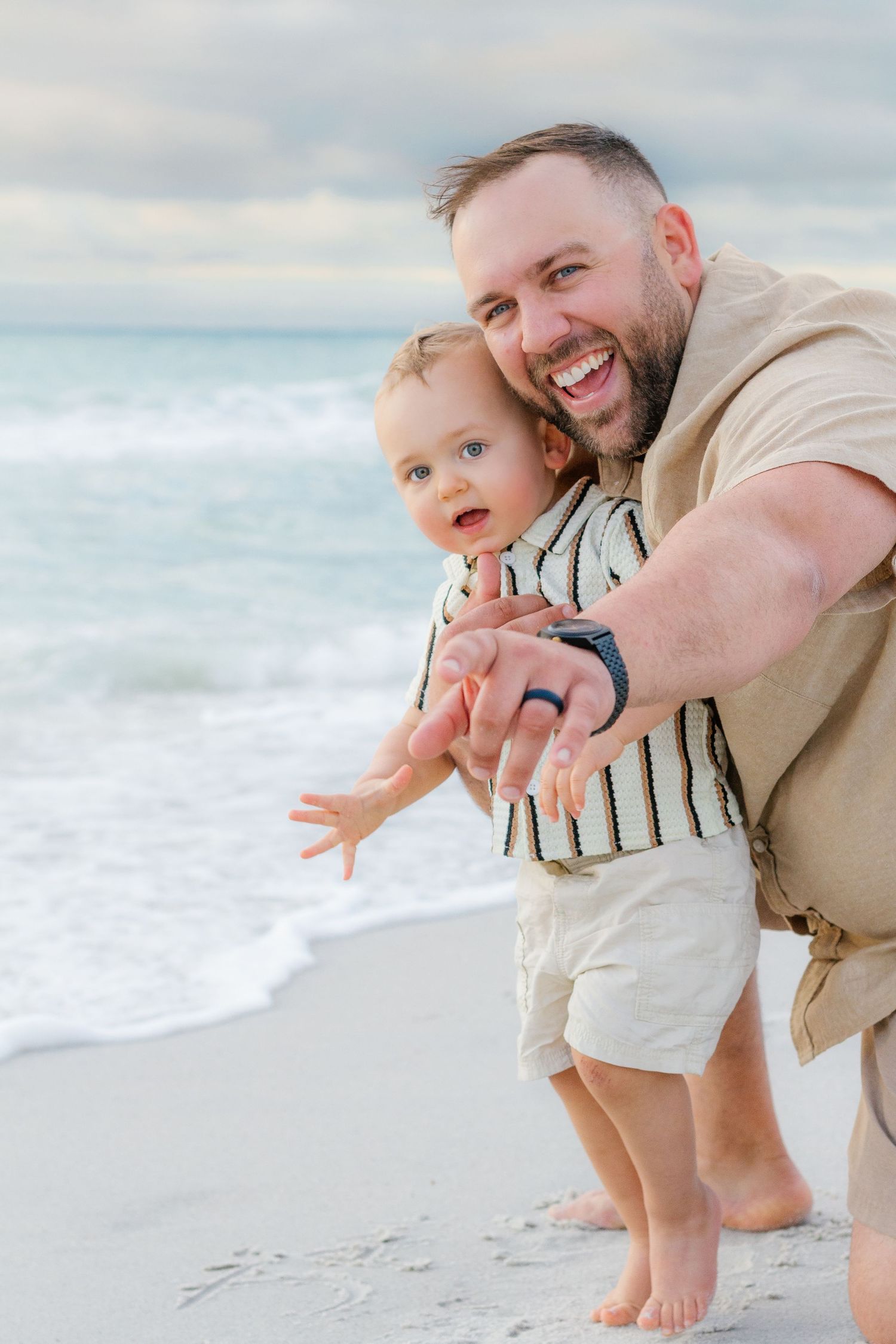 A laughing father helps his toddler son stand on a sandy beach with ocean waves in the background.