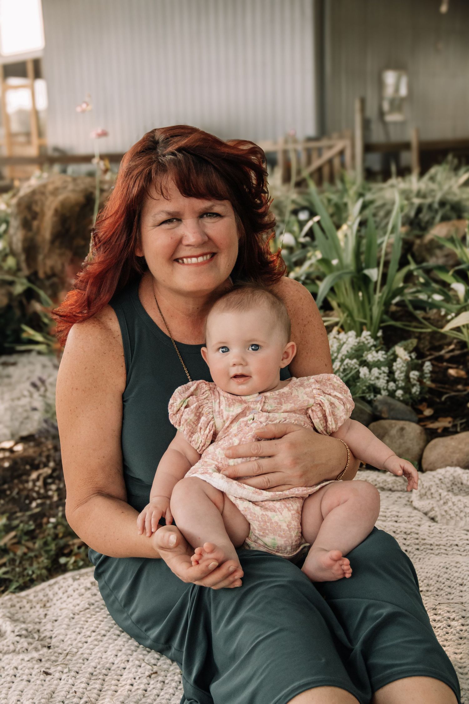Smiling woman with red hair holding a happy baby in a floral outfit, sitting outdoors near lush green plants.