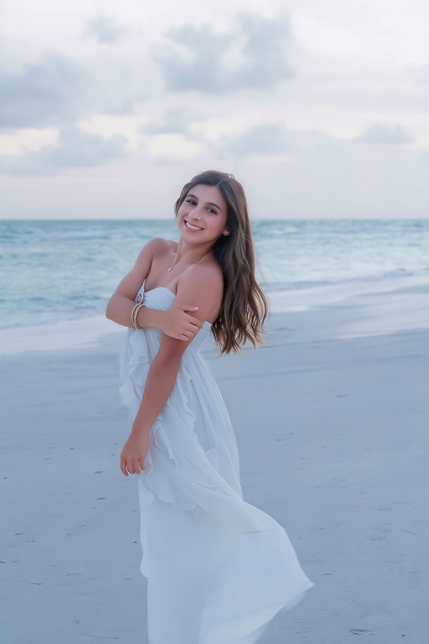Young woman in white dress smiling on a sandy beach with turquoise ocean waves in the background.