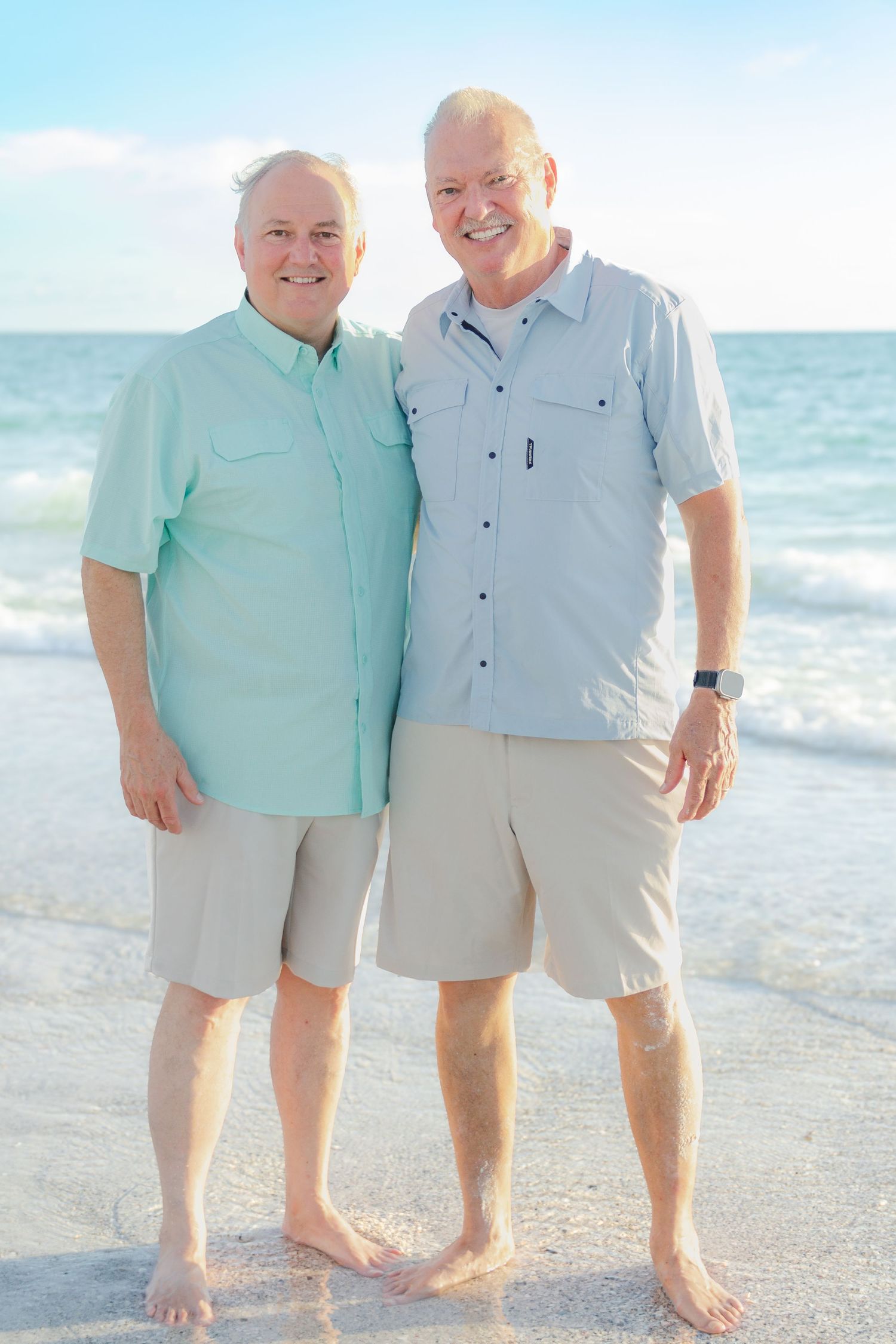 Two smiling senior men standing together barefoot on a sunny beach with ocean waves behind them.