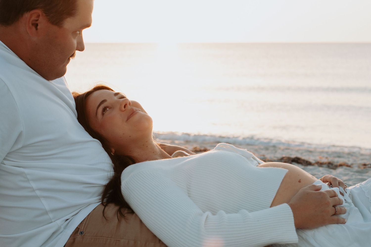 Couple relaxing on a beach at sunset, woman lying on man's lap in white outfits near the ocean.