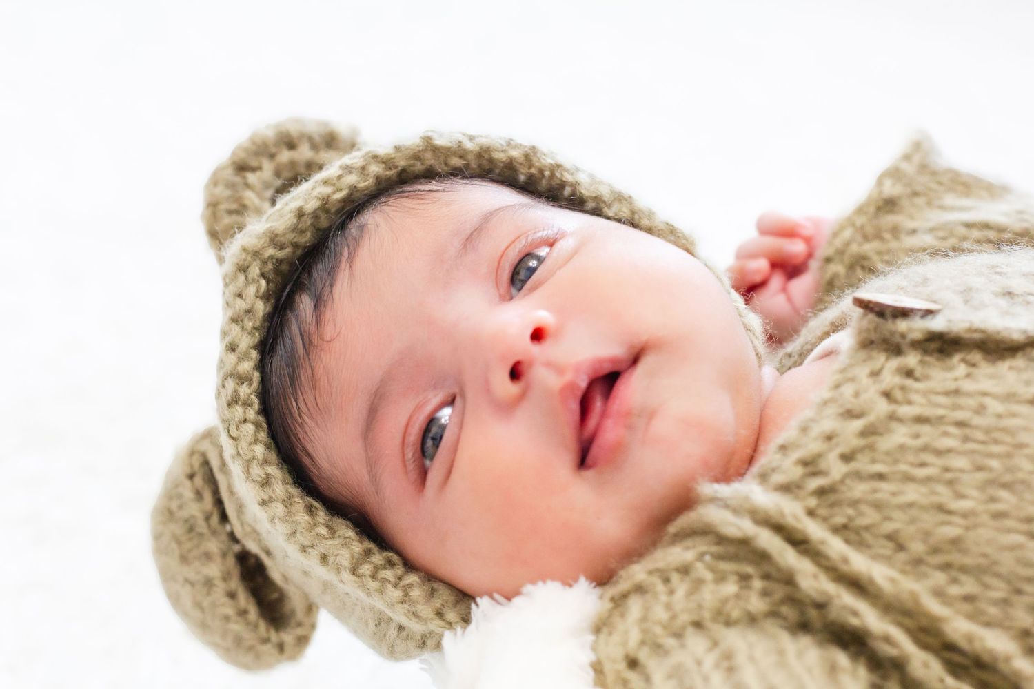 Smiling newborn baby wrapped in a cozy knit bear outfit with ears, lying on a soft white blanket.