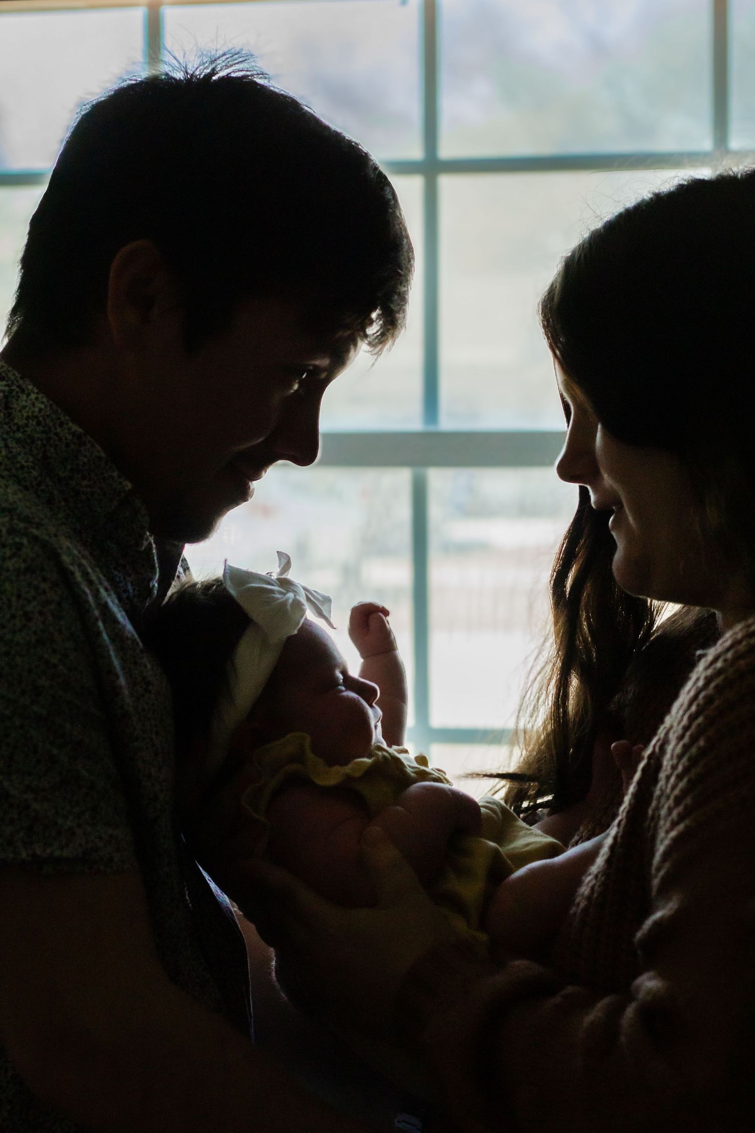 Silhouette of two parents lovingly gazing at their newborn baby by a bright window.