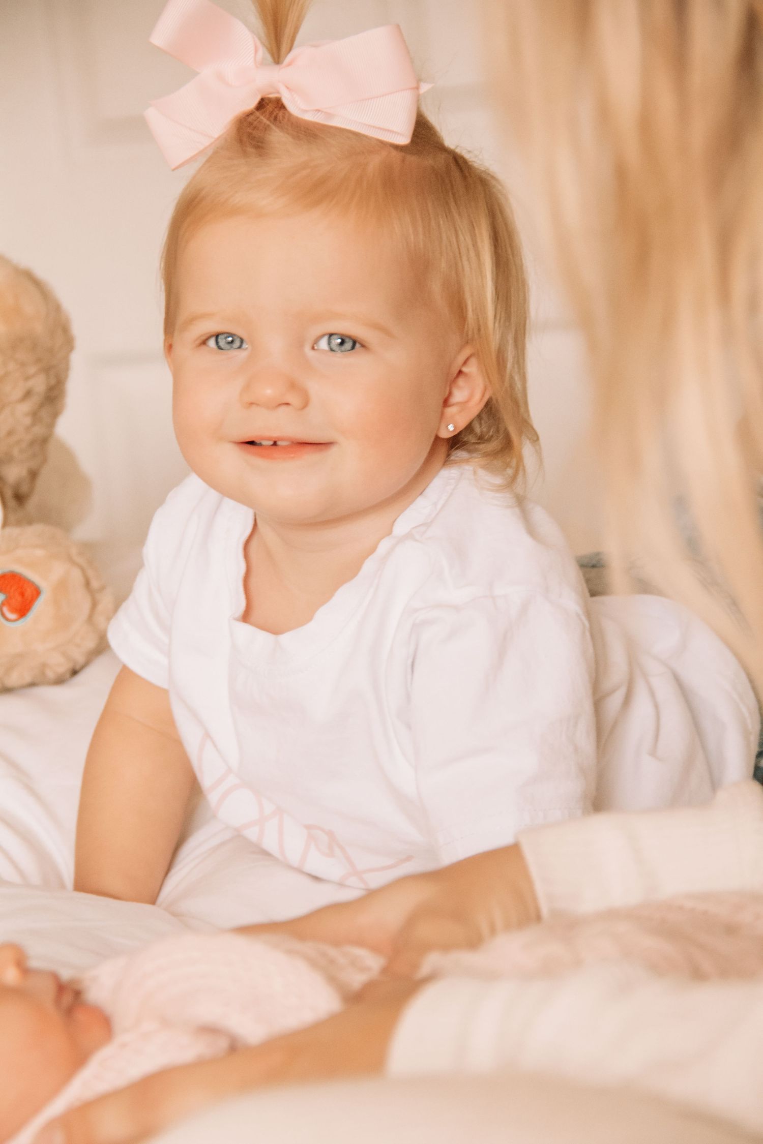 Adorable blonde toddler girl with blue eyes and pink bow smiles in white dress on bed with teddy bear.