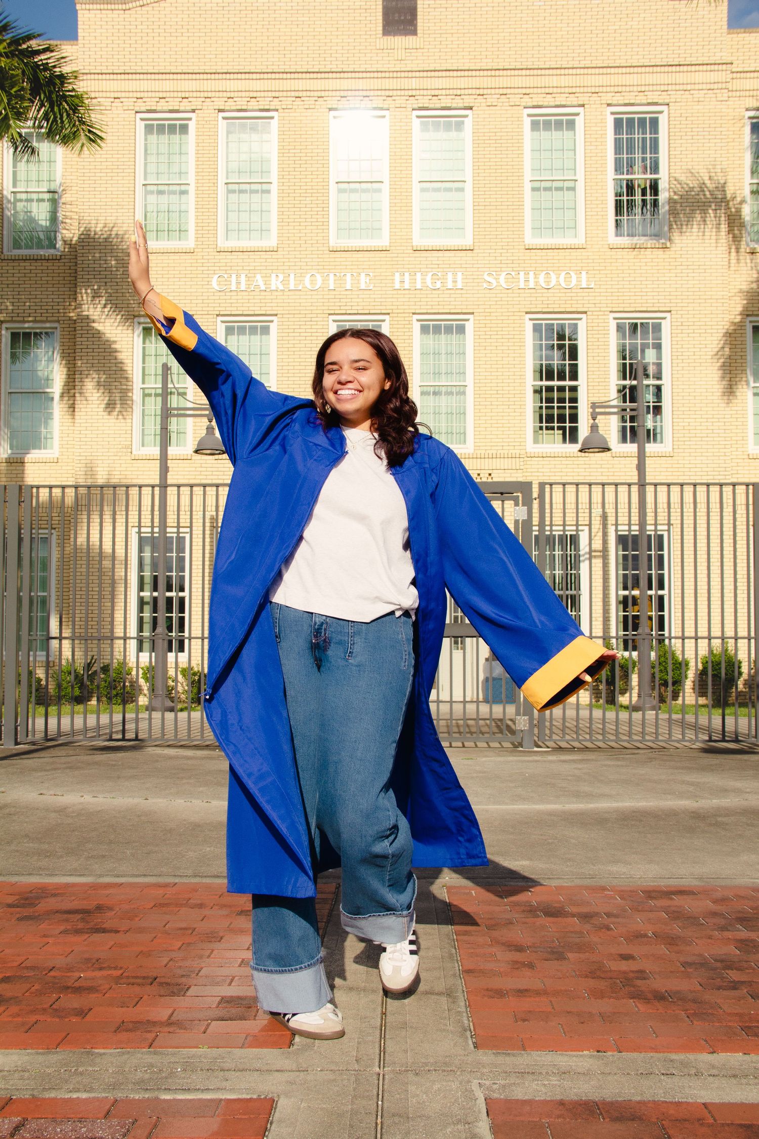 A smiling graduate in a blue and gold cap and gown poses in front of Charlotte High School on a sunny day.