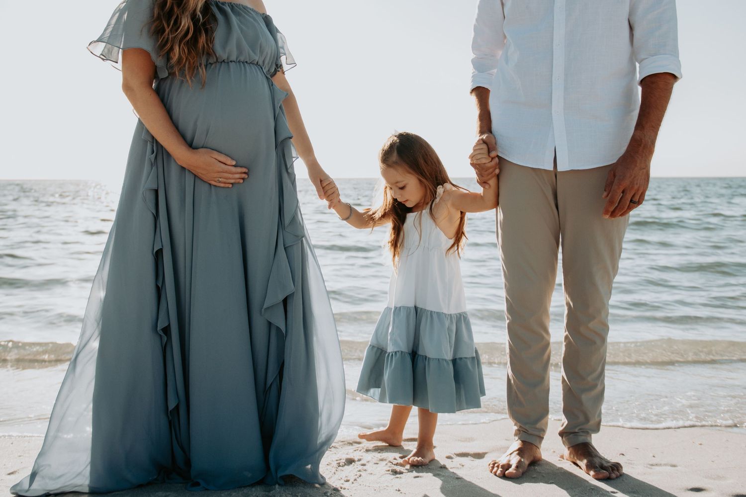 Pregnant woman in blue gown holding hands with young daughter and man on a sandy beach.