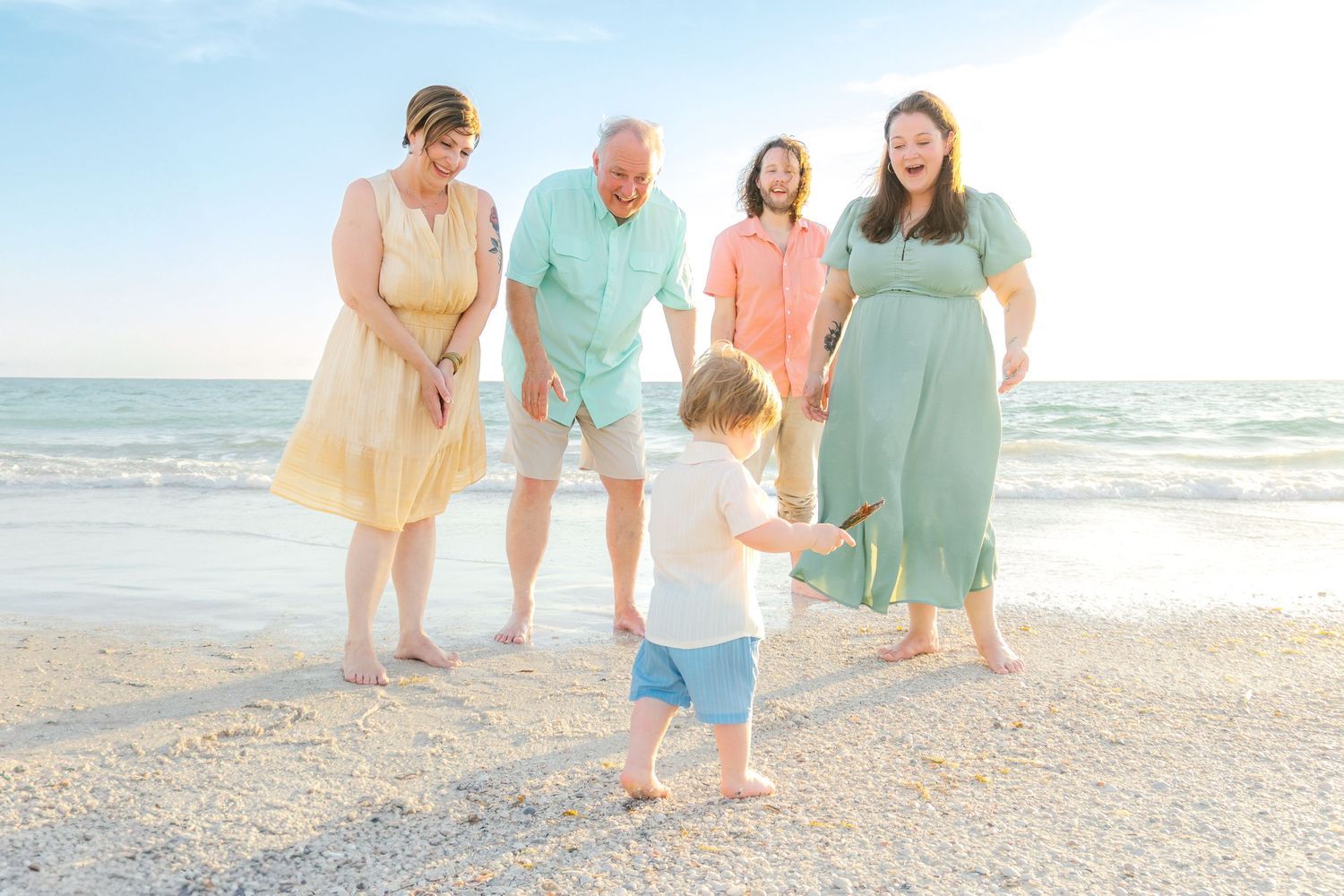 Family of five enjoying a sunny beach day, with a toddler taking first steps toward laughing adults in pastel outfits.