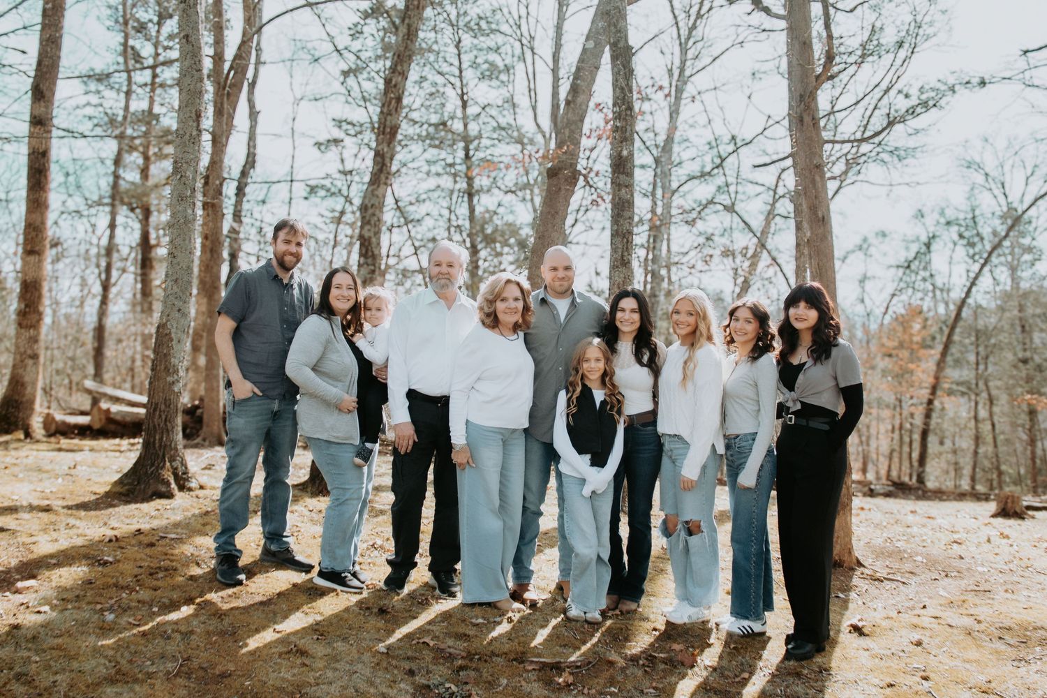 Large family group portrait outdoors in a wooded setting, posing together on a sunny day in casual coordinated outfits.