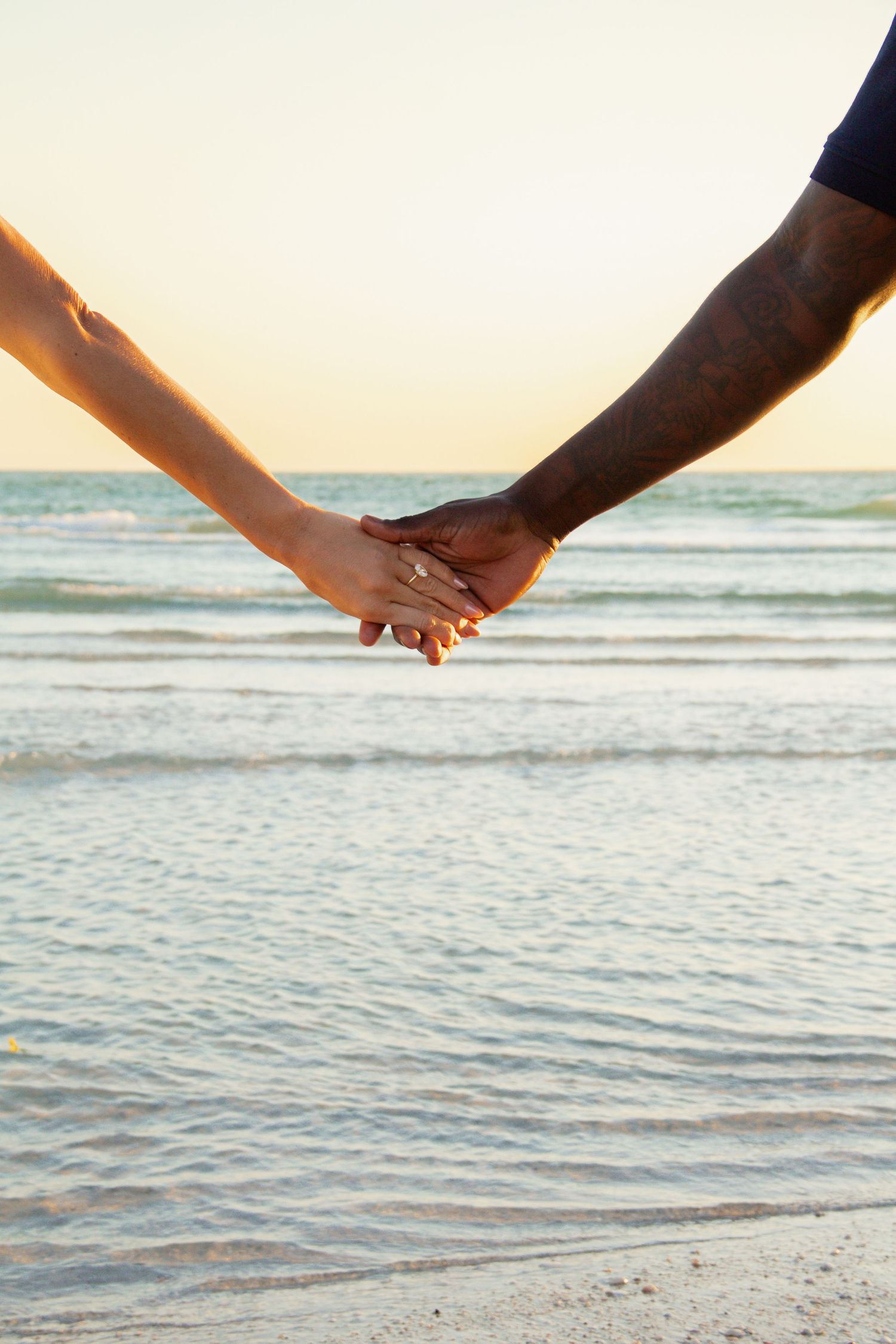 Interracial couple holding hands on the beach at sunset, woman wearing a diamond engagement ring.