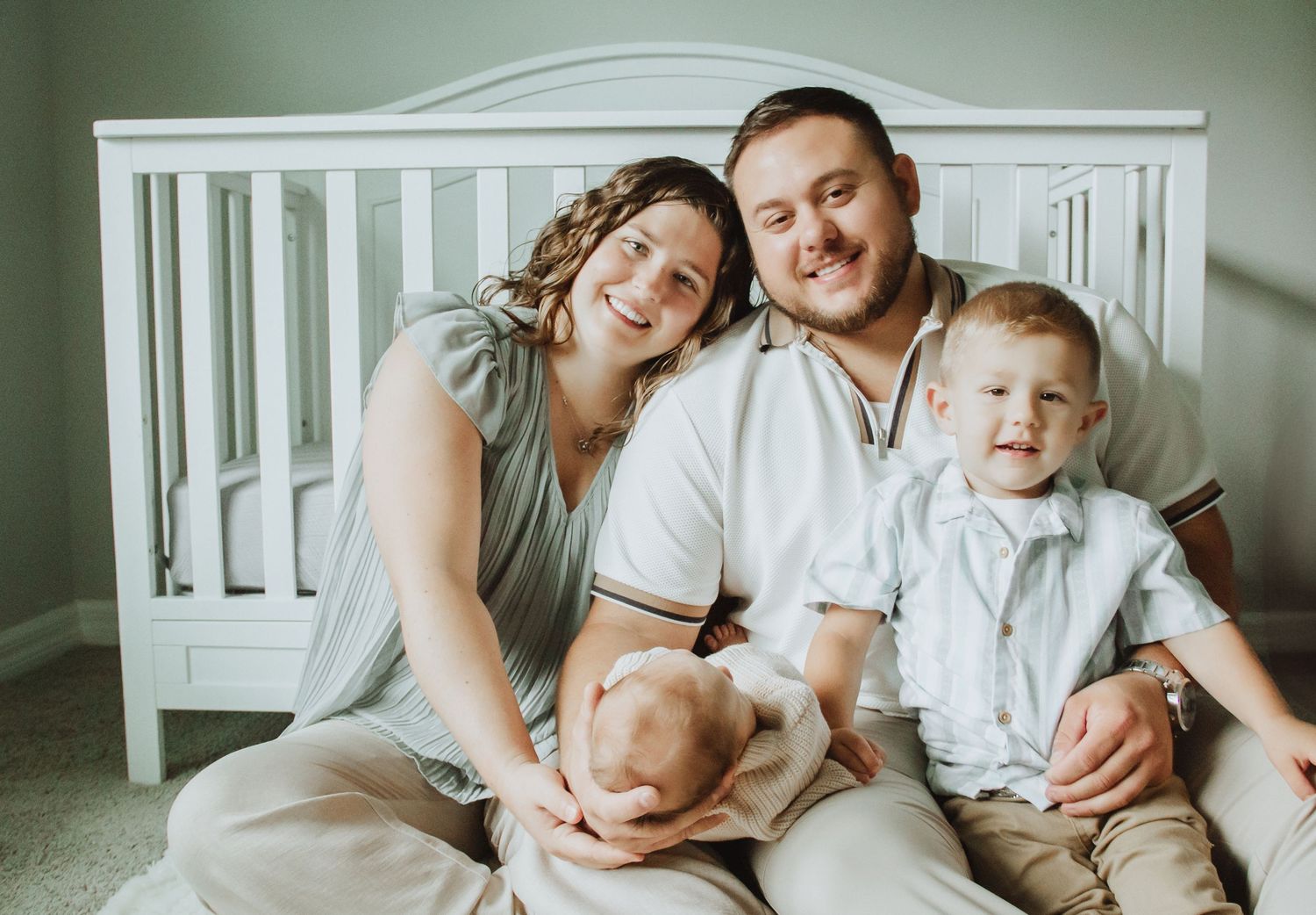 Happy family of four posing together near a white crib, parents smiling with newborn baby and toddler son.