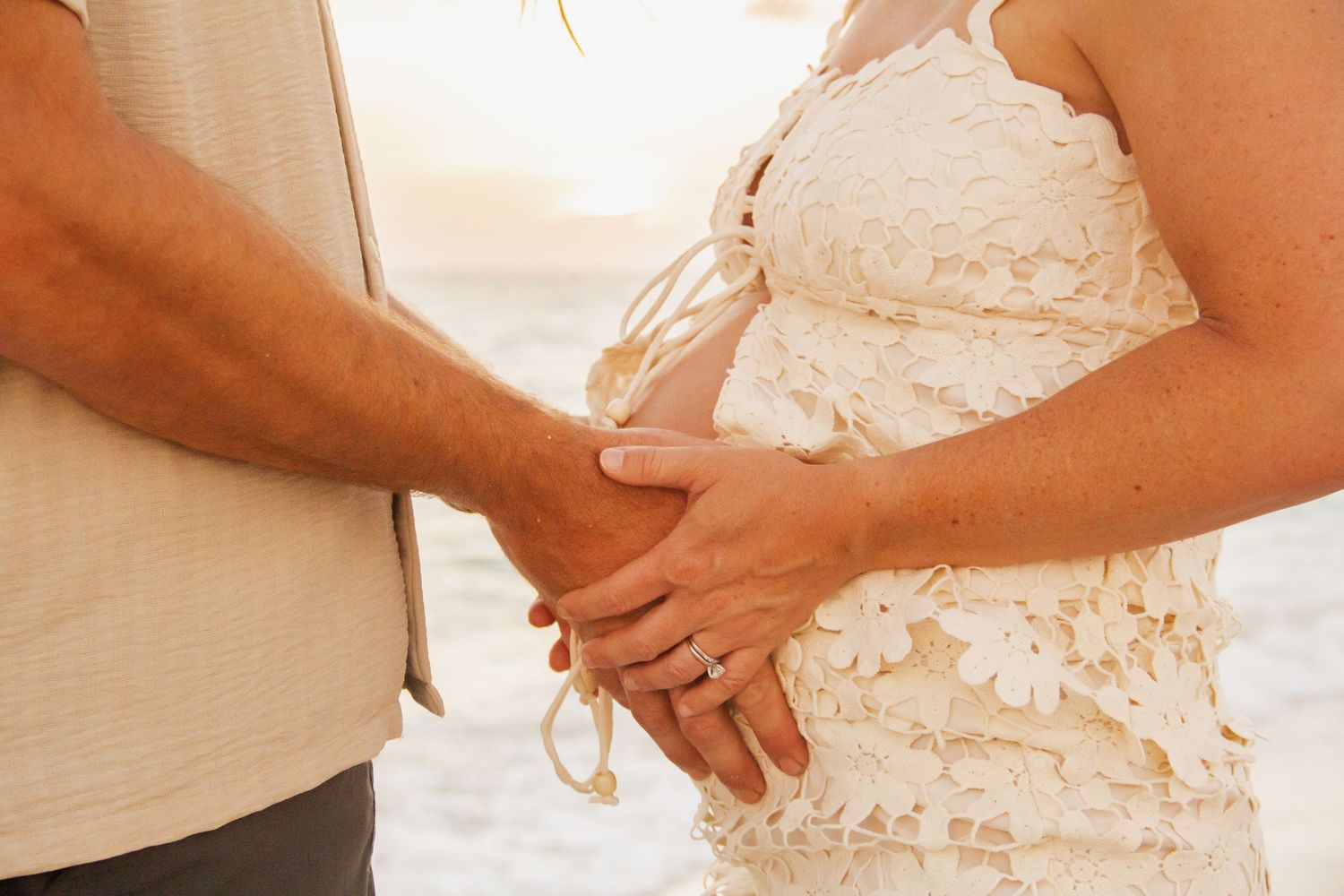 Couple holding hands on beach, pregnant woman in white lace dress with engagement ring, romantic maternity photo.