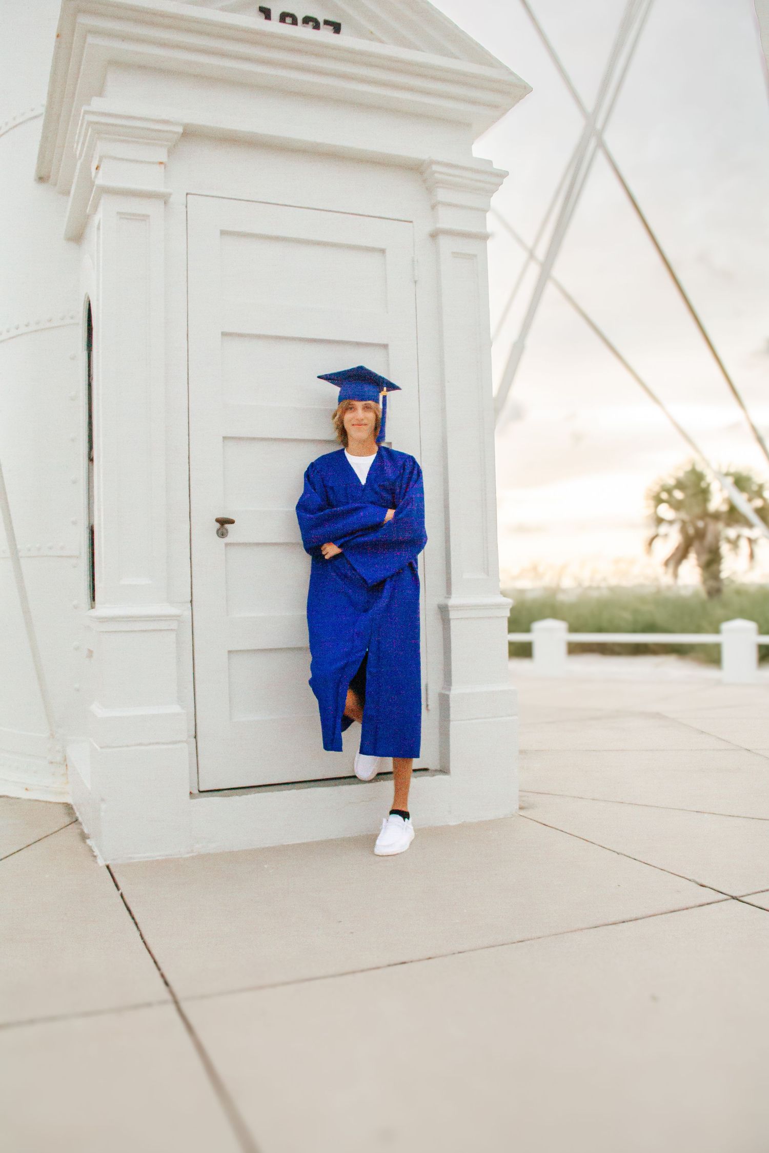 Graduate in blue cap and gown leaning against white building door, arms crossed, at sunset.