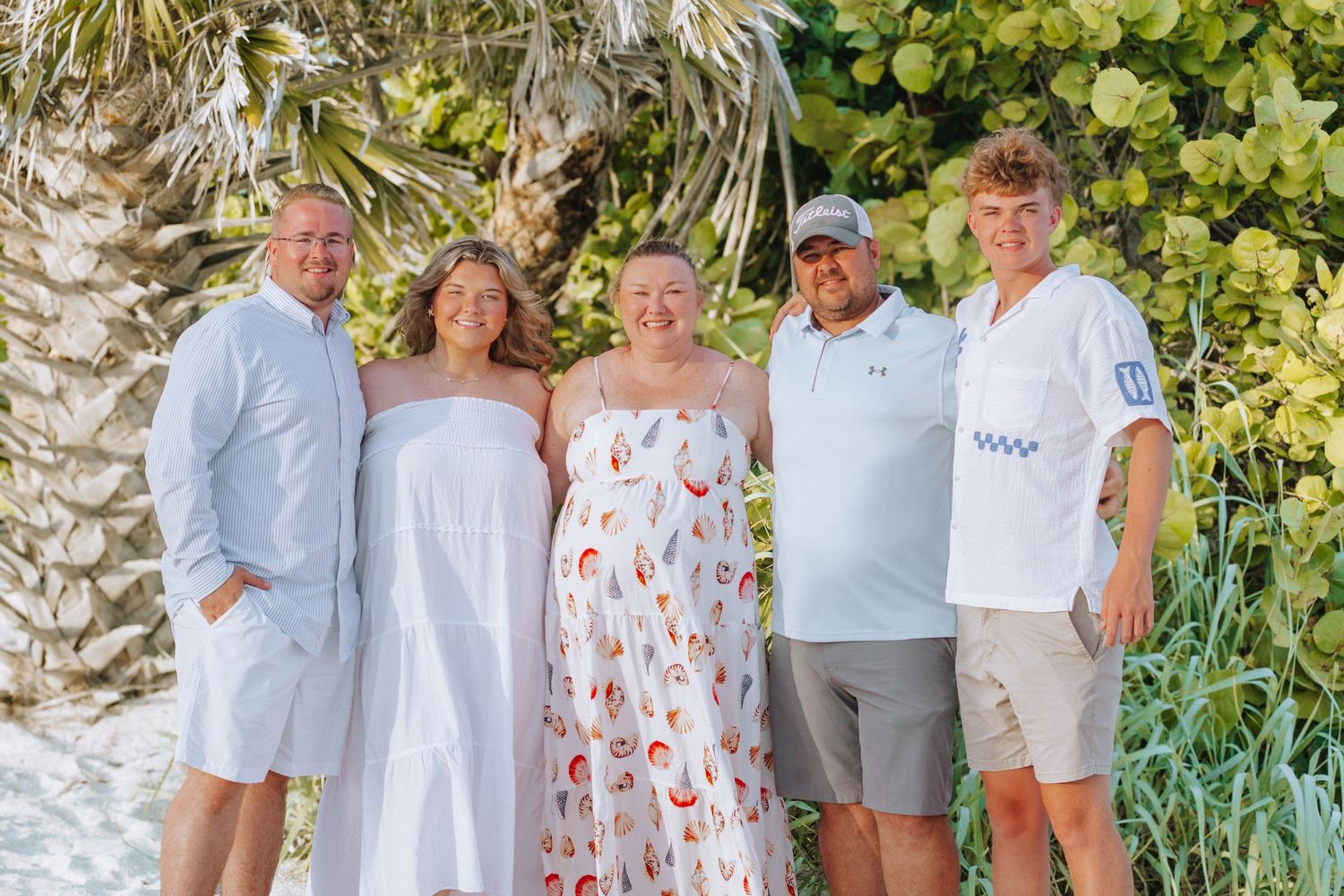Five smiling people pose together on a sandy beach surrounded by lush tropical greenery and a palm tree.