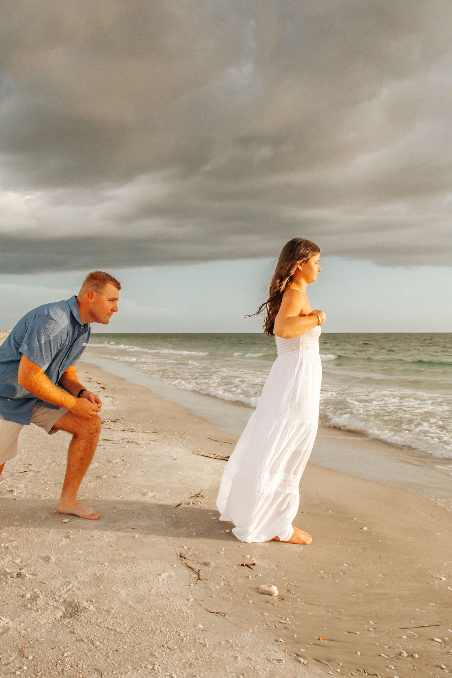 Couple on a beach at sunset, man kneeling as woman in white dress stands by the shoreline under cloudy skies.