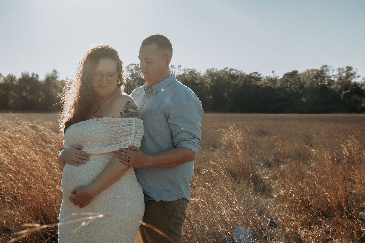 Couple in golden field, pregnant woman in white lace dress, man in blue shirt embracing her bump, backlit by warm sunlight.