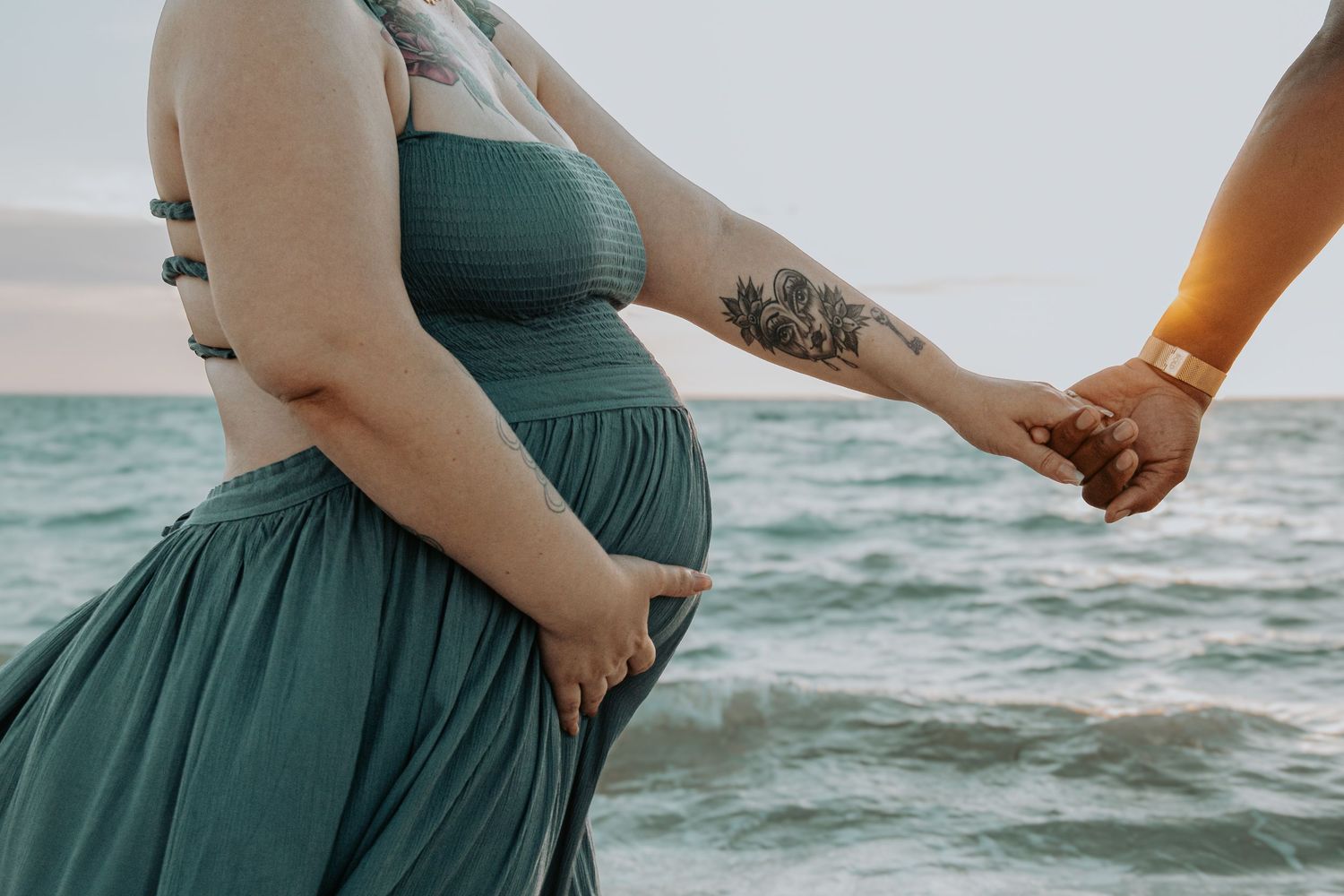 Pregnant woman in teal dress holding hands with partner on beach at sunset.