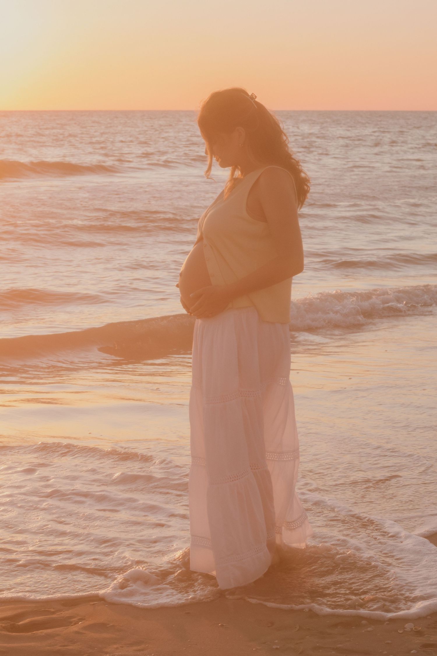 Pregnant woman in white dress standing on beach at sunset, cradling her baby bump by the ocean waves.